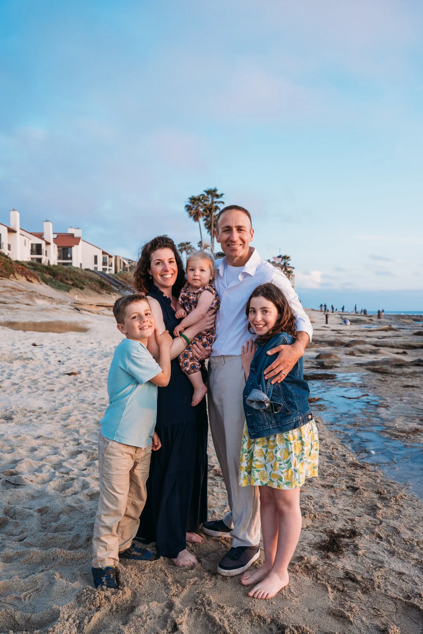 A family of five standing together on a beach at sunset, with houses and palm trees in the background. Alisha Mowry Photography Military Photographer San Diego