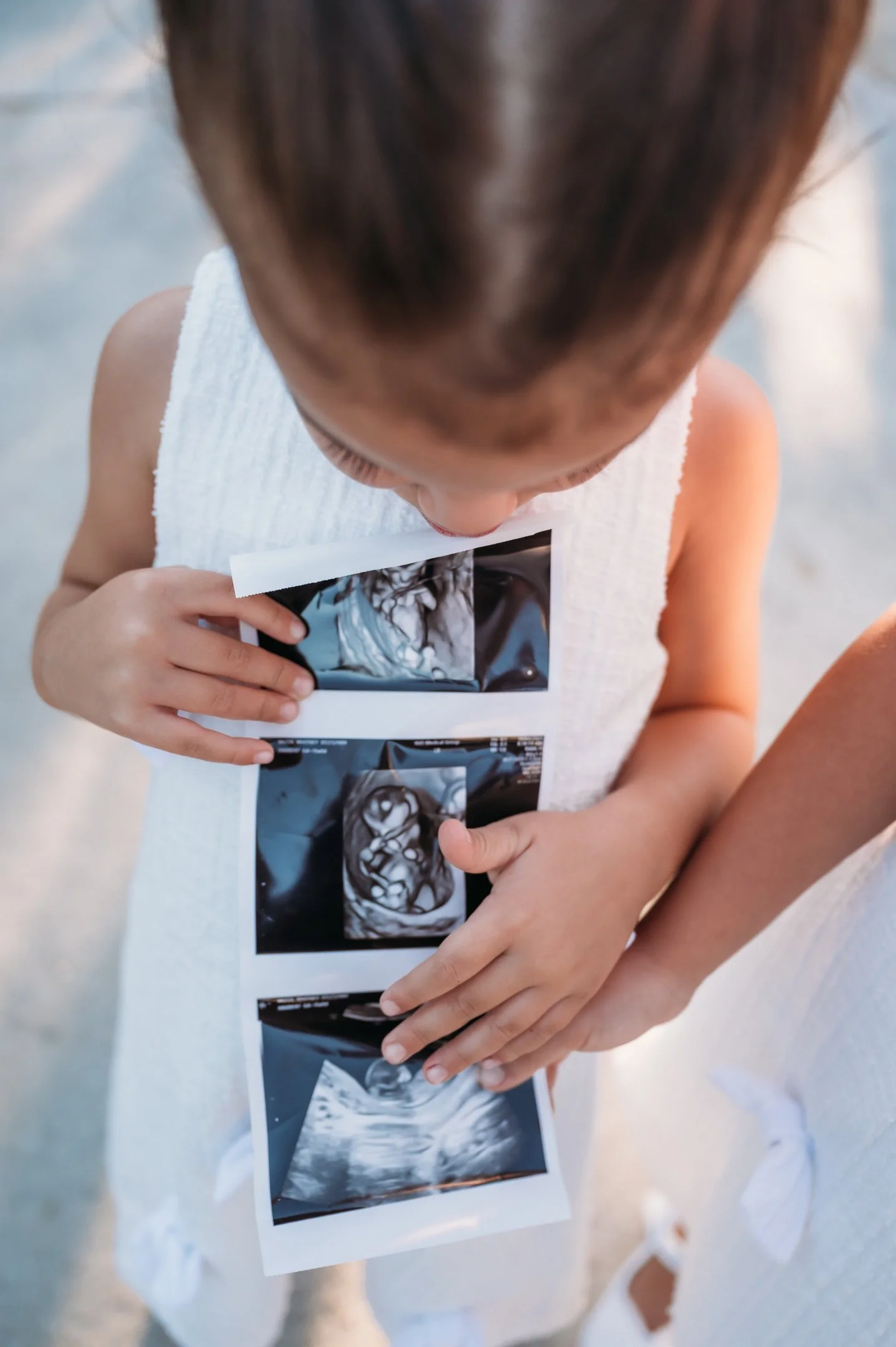 A young child holding and looking at ultrasound photos, with a beach background. Alisha Mowry Photography Military, Brand, and Portrait Photographer San Diego CA