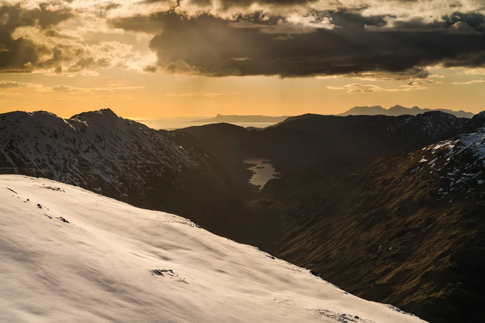 “My wintery perch overlooked Loch Beoraid, its blank headwall lost in matt shadows. Above that inky blackness, an orange fire reigned over Eigg and the Skye Cuillin.”