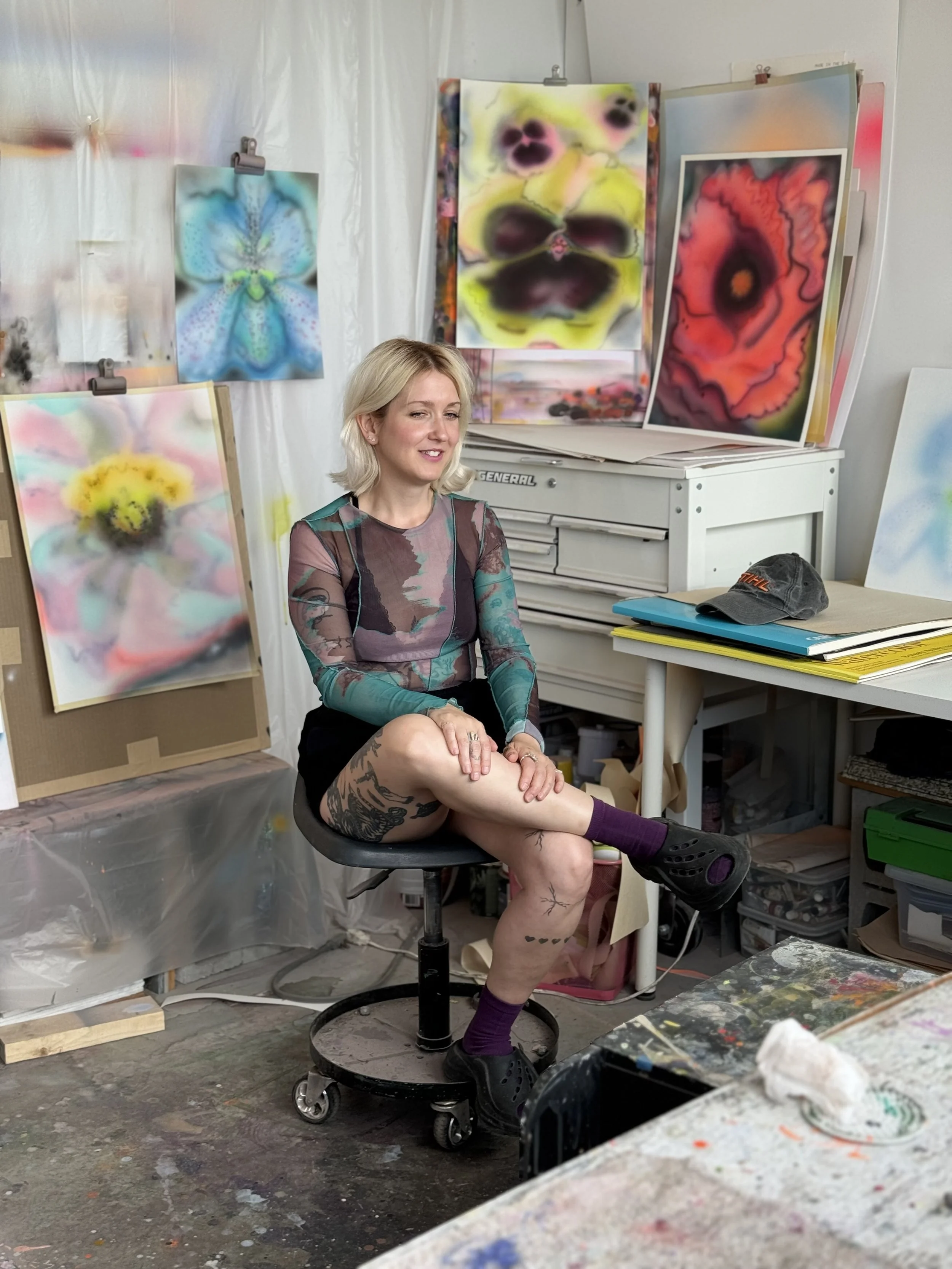 A woman with blonde hair sitting on a stool in an art studio surrounded by colorful abstract paintings, with painting supplies and artwork visible around her.