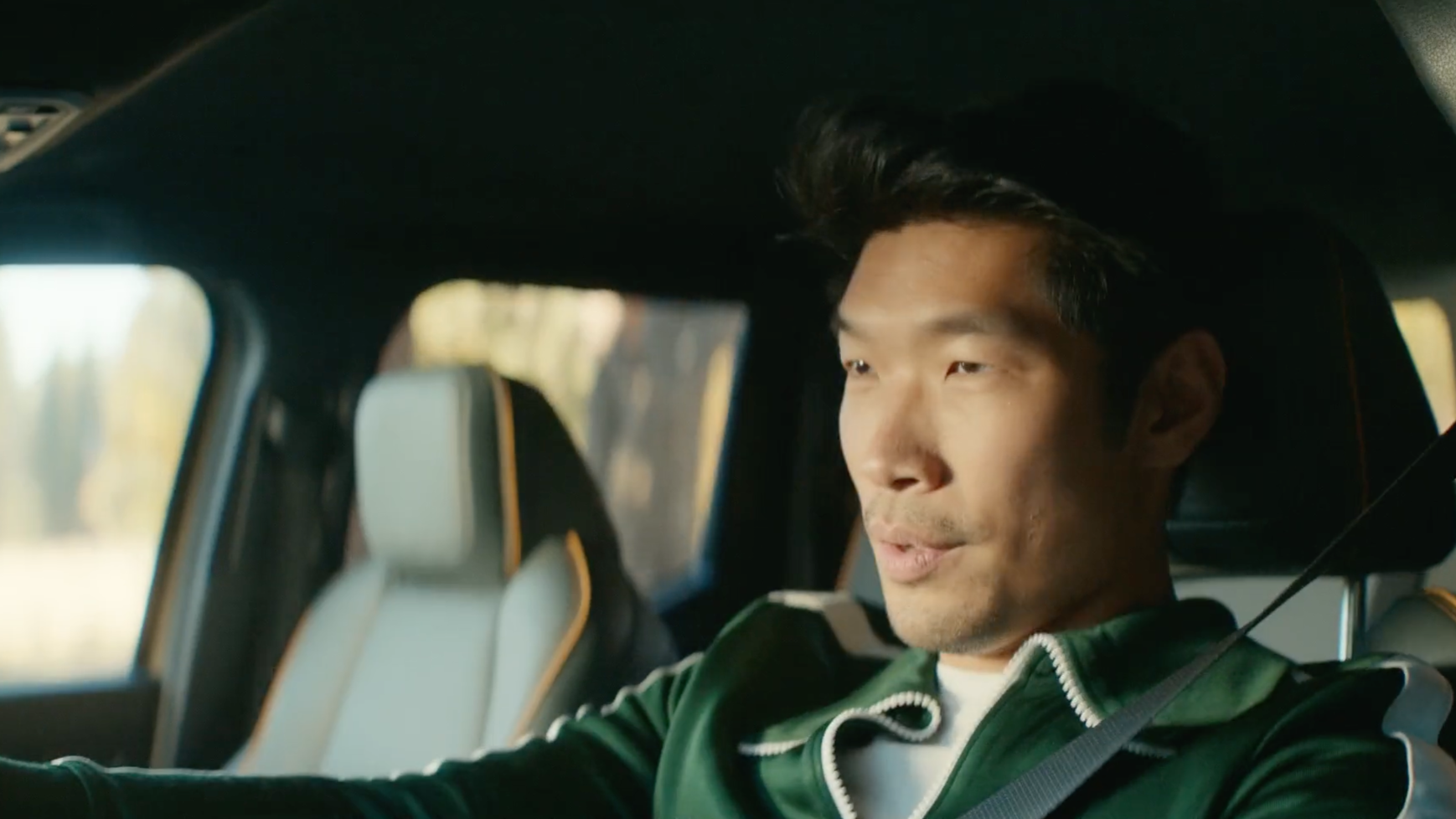 A man with black hair, wearing a green jacket, sitting in a car and talking.