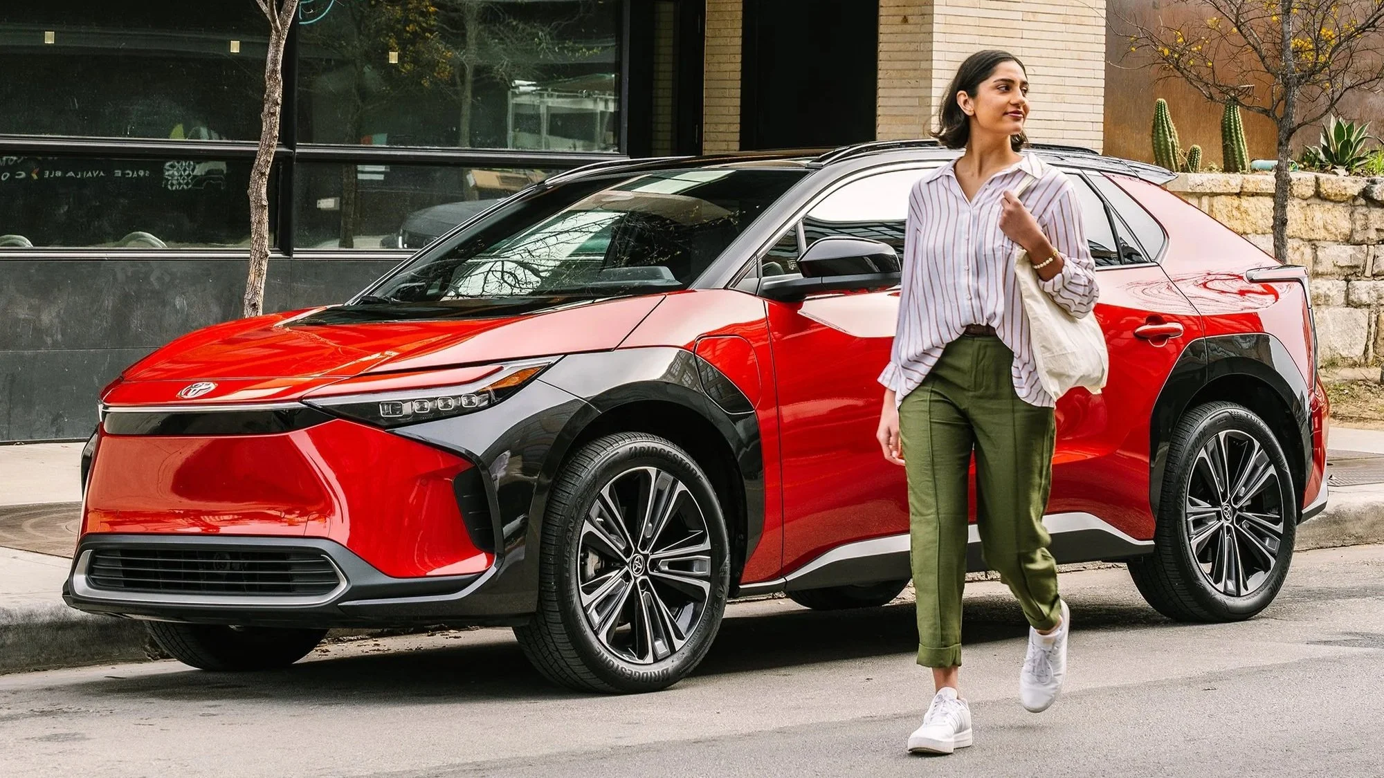 A woman walking on a city street past a red and black Toyota electric vehicle, with a background of a storefront, trees, and cacti.