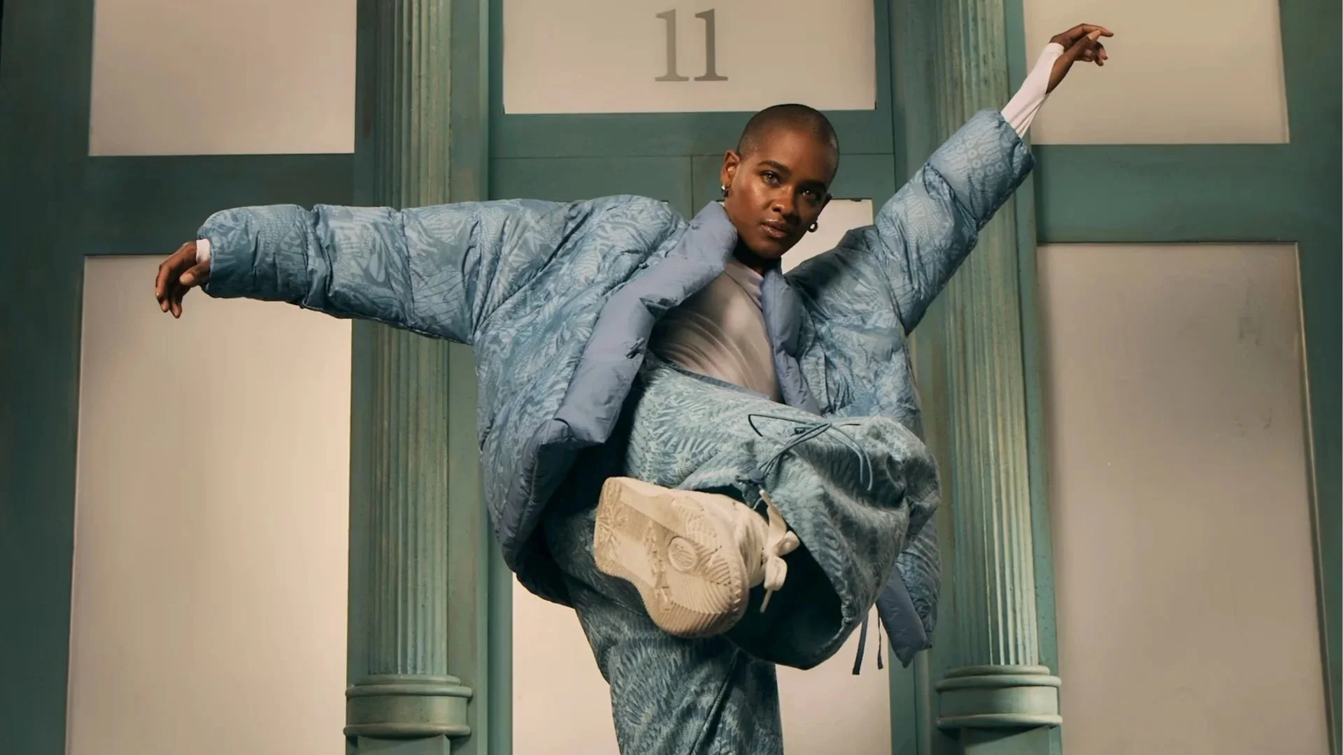 A woman with a shaved head wearing a blue patterned puffer jacket, matching pants, and sneakers, raising her arms in a dance pose in front of a teal-colored wall with columns.