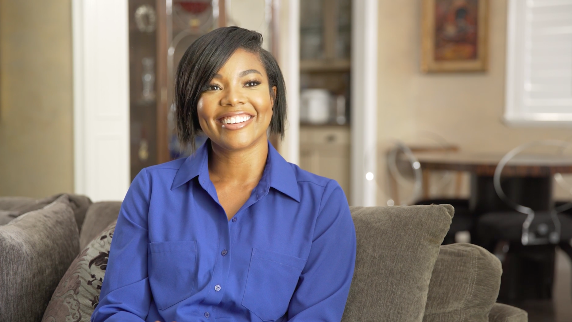 A woman sitting on a sofa, smiling, wearing a blue button-up shirt in a cozy living room.