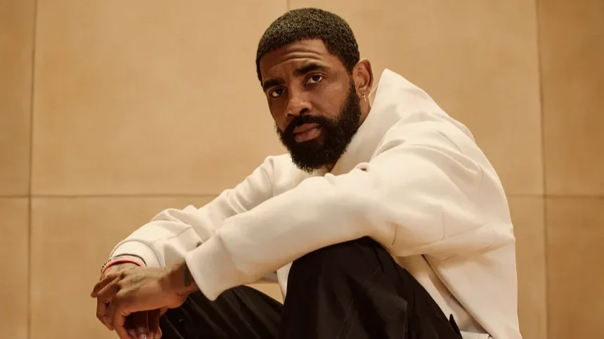 A man sitting on the floor, wearing a white hoodie and black pants, with a serious expression, against a plain light brown background.