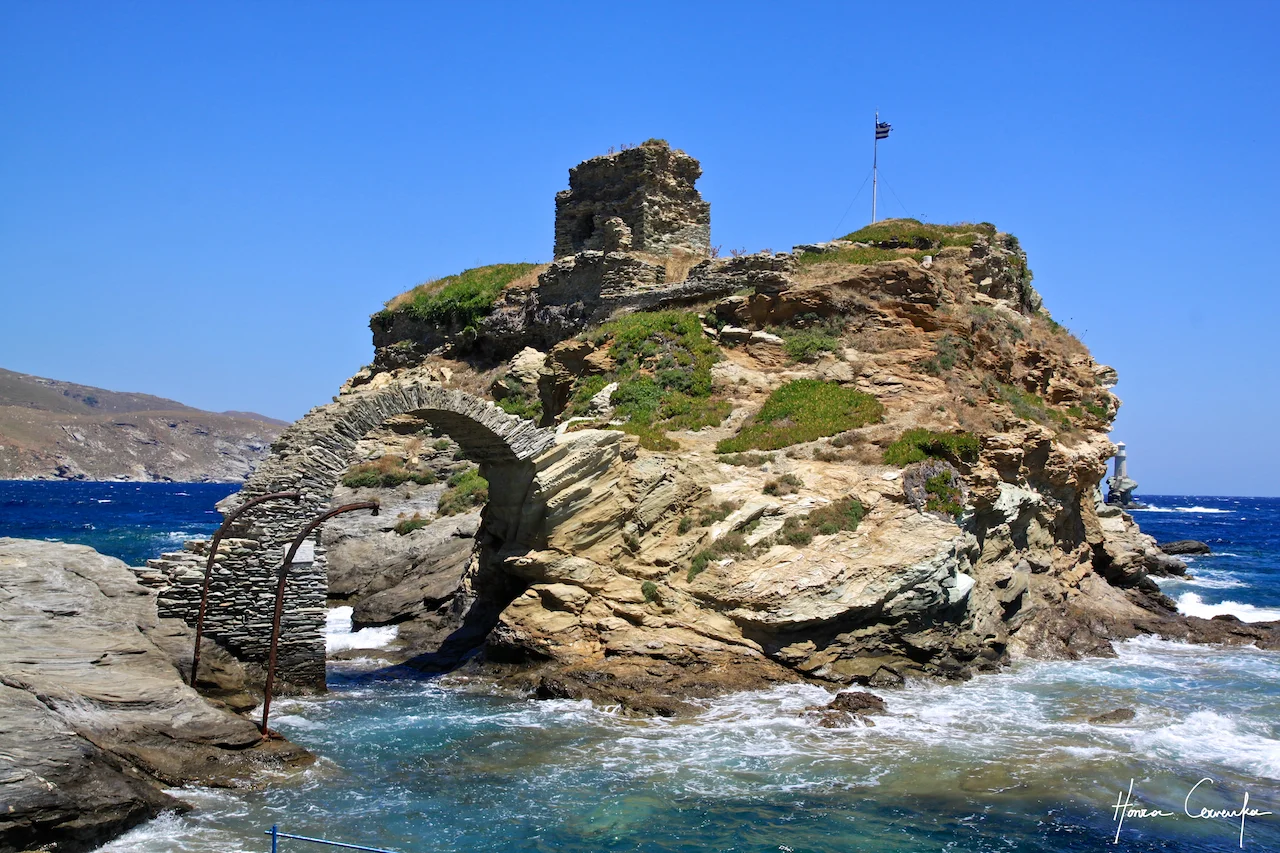 The two "lampposts" that you see are actually remnants of old cranes. In the winter, when the Meltemi subsides significantly (to the point of being virtually non-existent), the sea is calm enough for small boats to pass under the bridge and unload. I cannot imagine it has been used in the past few centuries, but there it is. In the summer, however, the passage would have been suicidal--any ship would end up on the rocks with these waves.