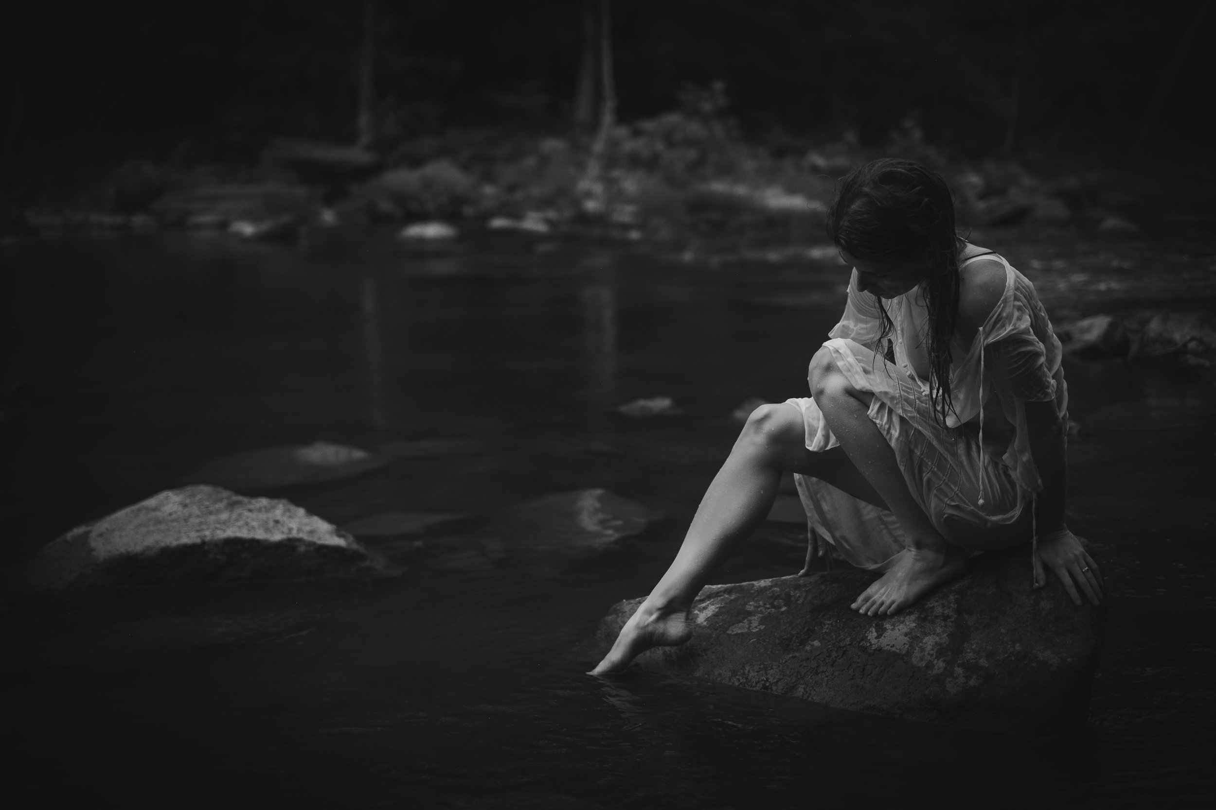 woman wearing sheer dress sitting on a rock dipping toe in the river