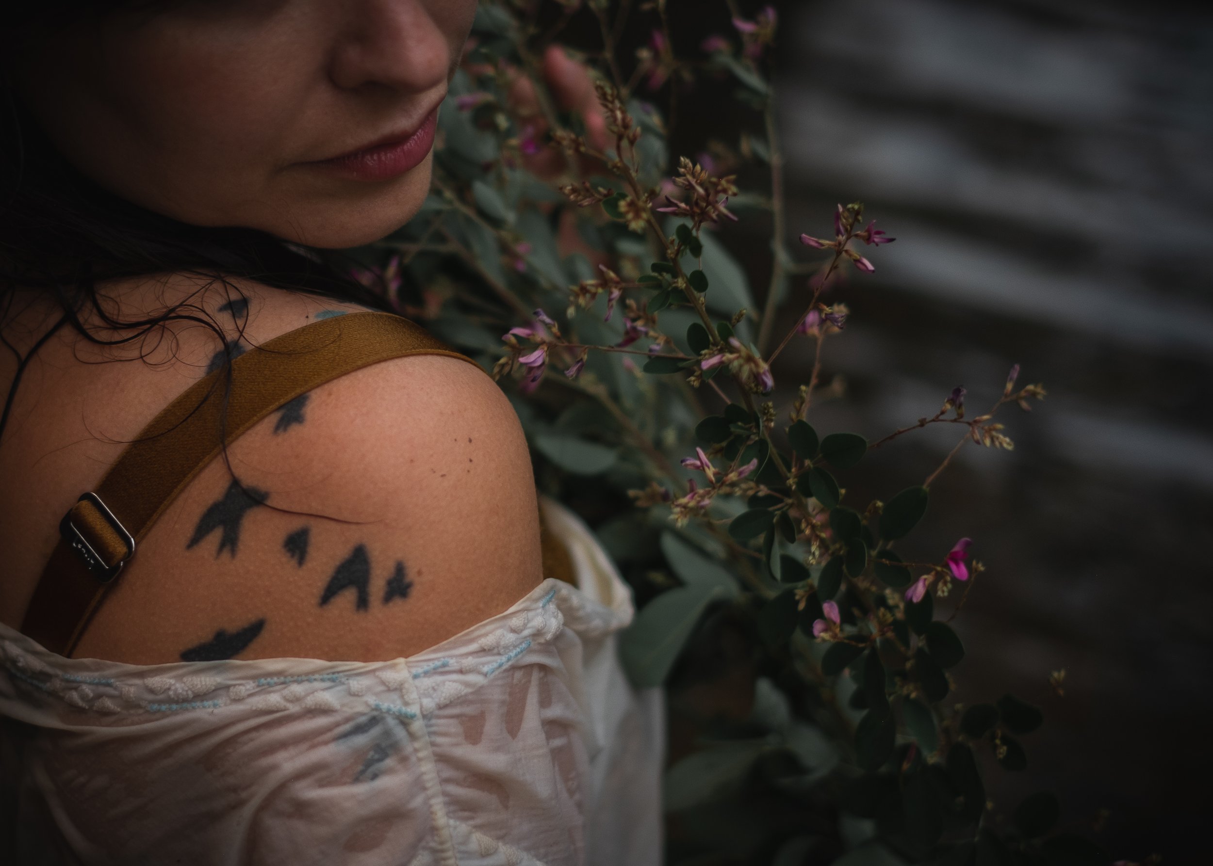 woman's tattooed shoulder of birds wearing a sheer wet dress holding wild purple blooms and branches