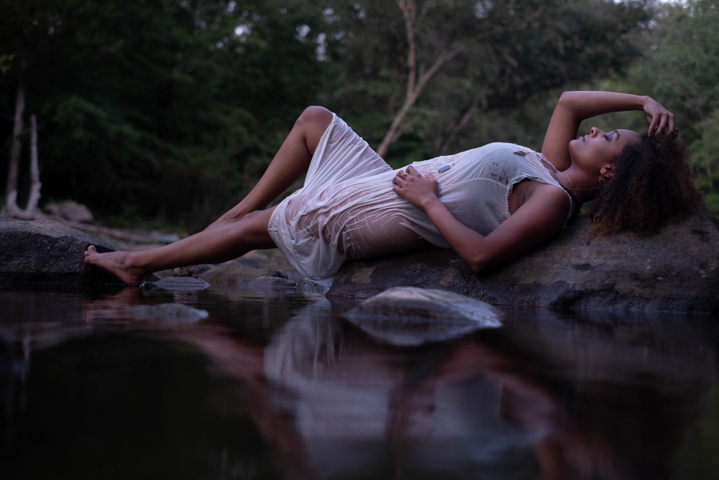 Outdoor boudoir of a woman with curly ethnic hair laying on top of rocks in river wearing Jennifer Dawes Jewelry