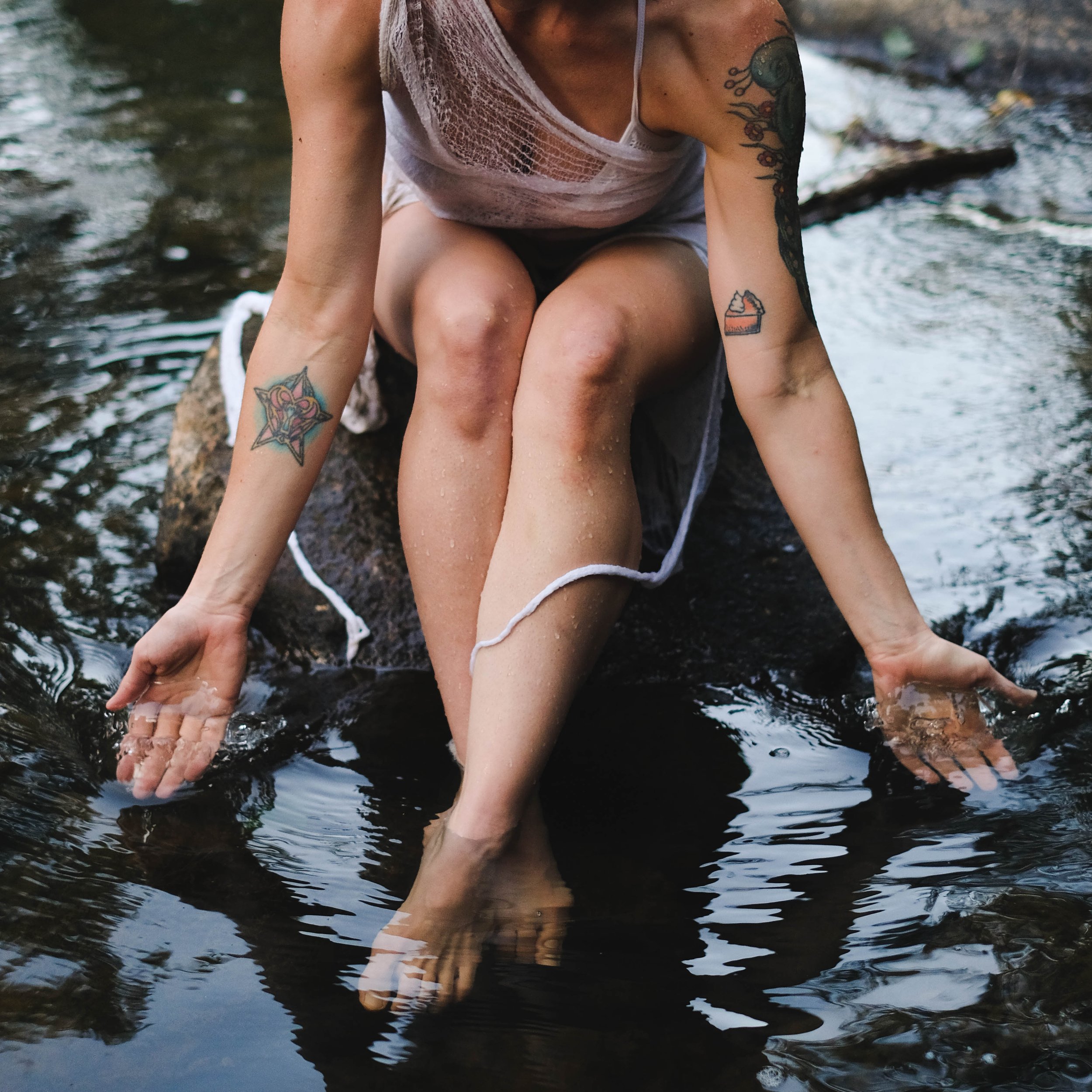 tattooed woman sitting on rock in a river with legs crossed and dipping hands into the water