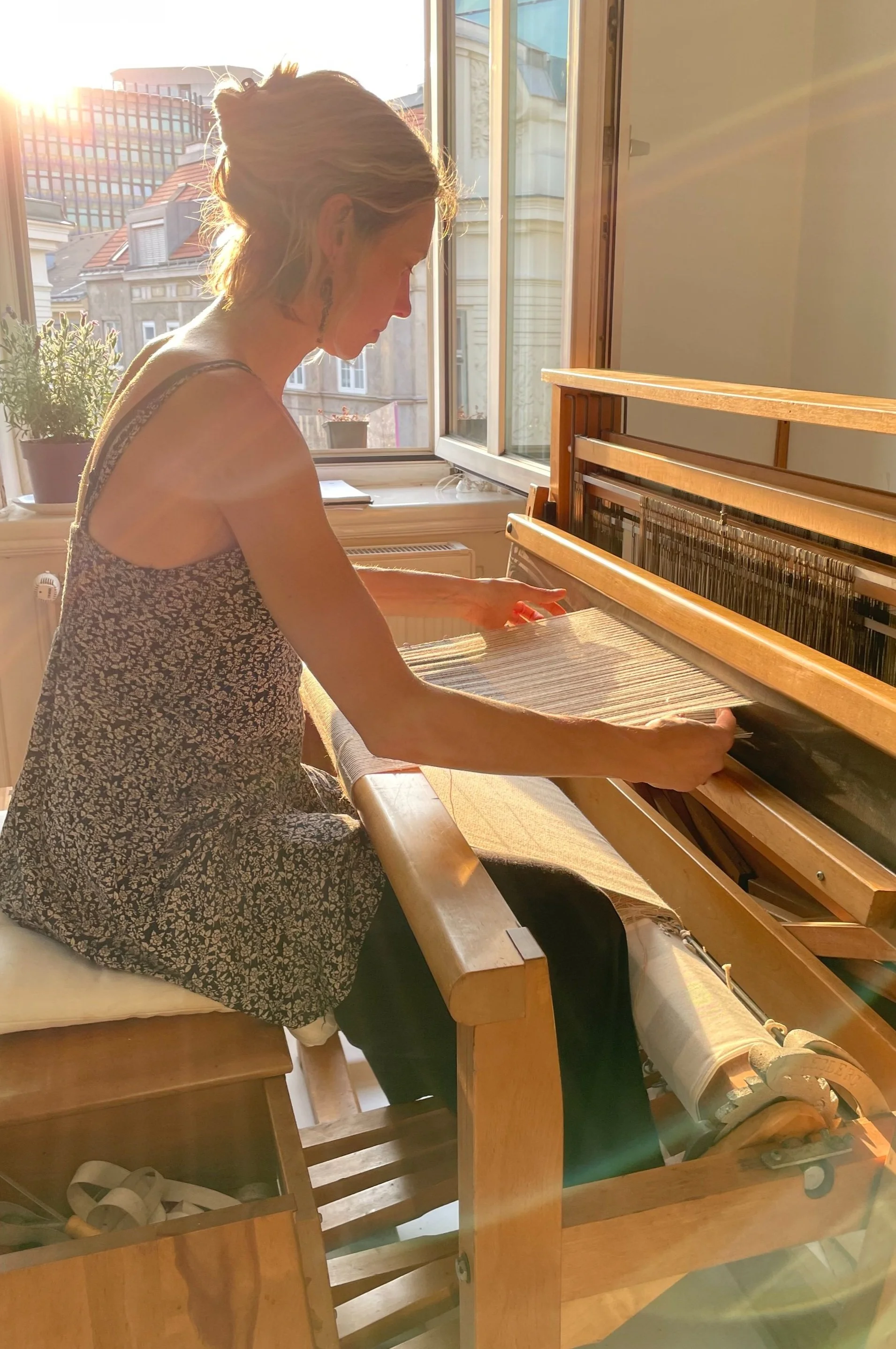 A woman sitting at a wooden loom, weaving fabric near a window with sunlight streaming in. Outside, buildings with red roofs and a modern glass building are visible.