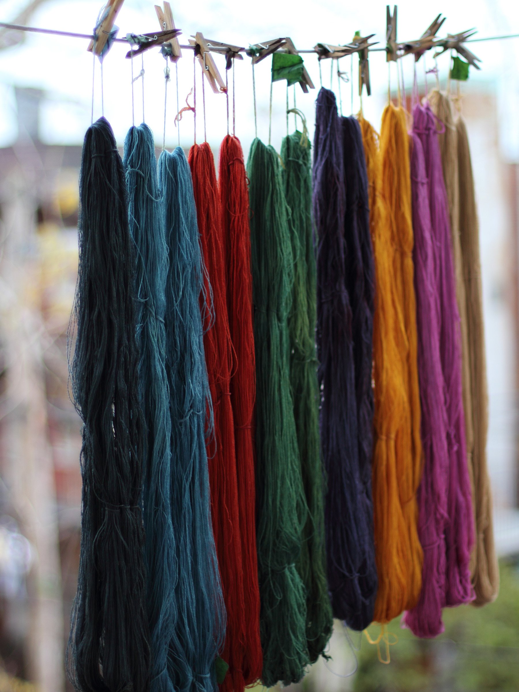 Multiple colorful skeins of yarn hanging on a clothesline with clothespins, outdoors with blurred background