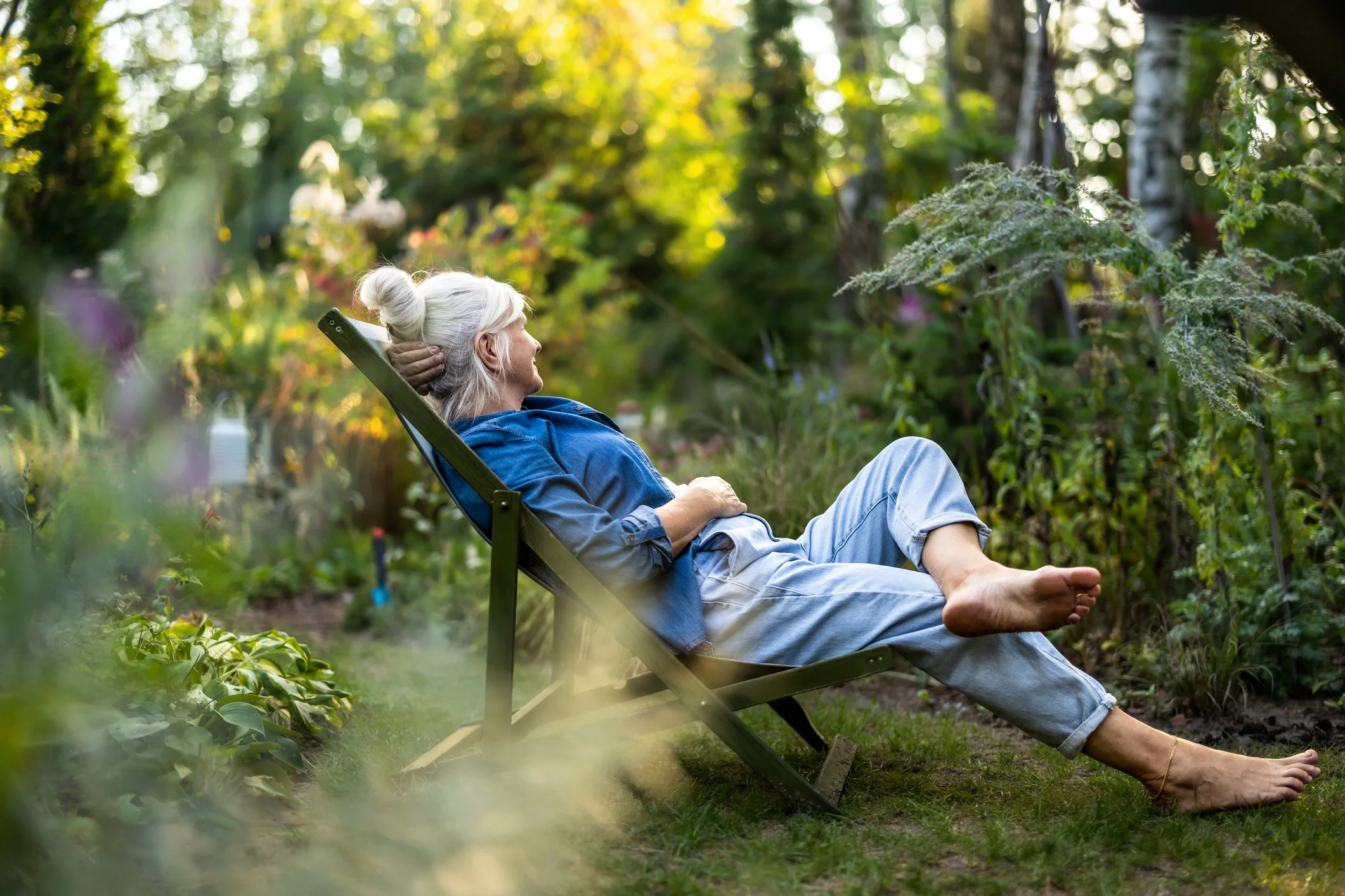 An elderly woman with gray hair in a bun relaxing on a garden lounge chair in a lush green garden, lying back with her hands behind her head and legs extended, enjoying the peaceful outdoor setting.