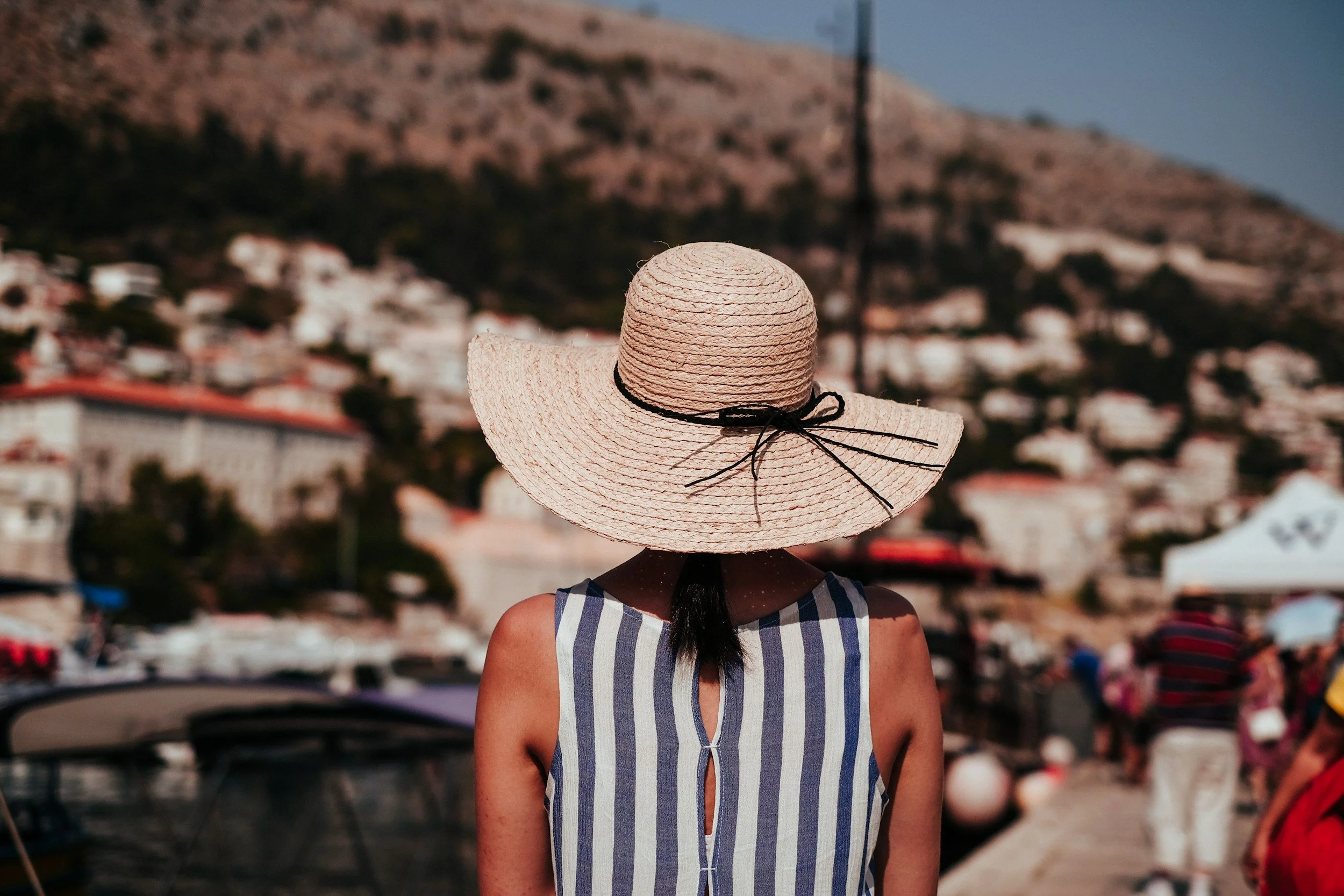 A woman in a striped sleeveless top and wide-brimmed straw hat with a black ribbon, standing at a marina with boats, on a sunny day, with a hillside town in the background.