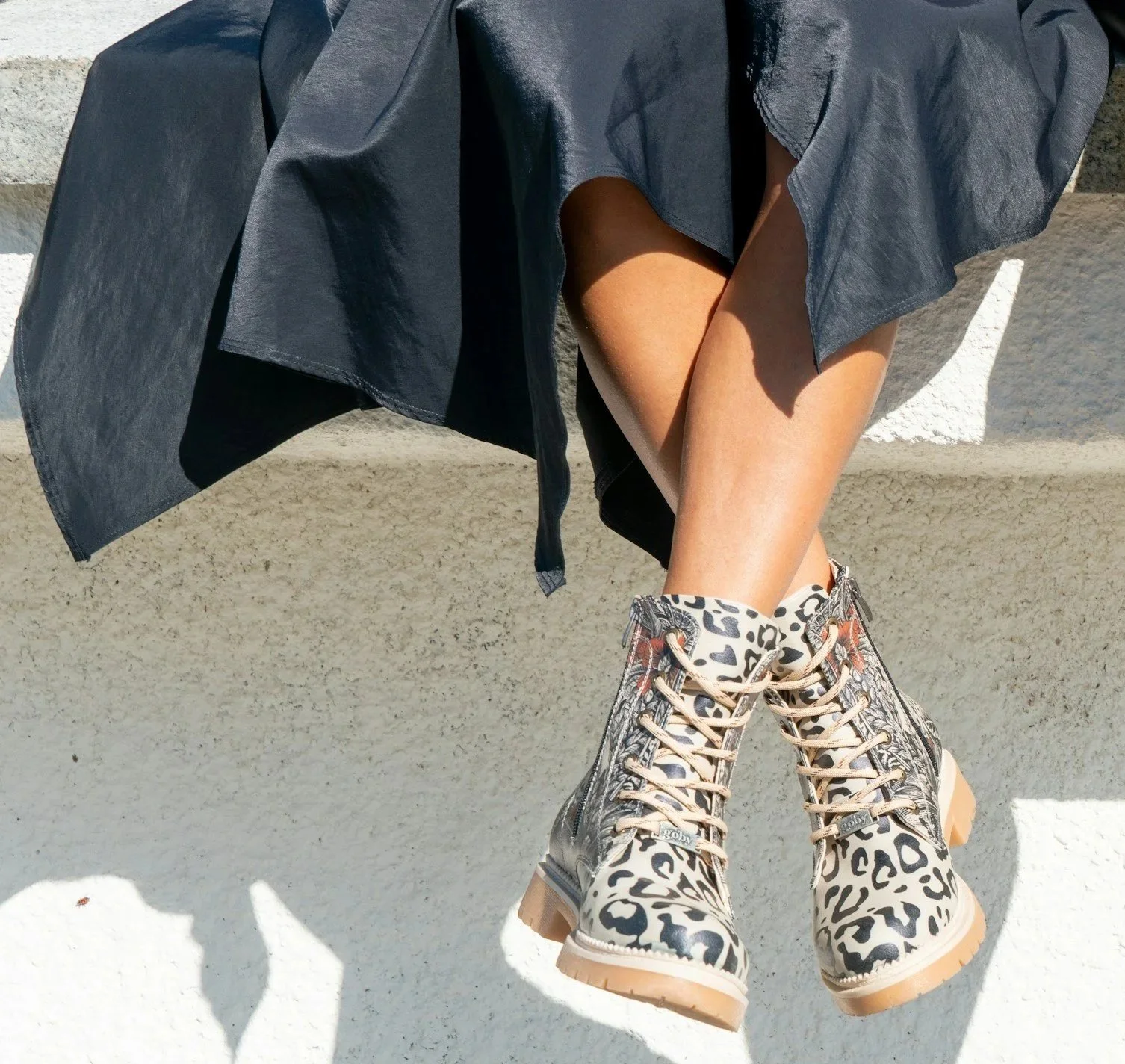 Person sitting with legs crossed, wearing beige leopard print boots and a dark skirt or dress, against a textured wall background.