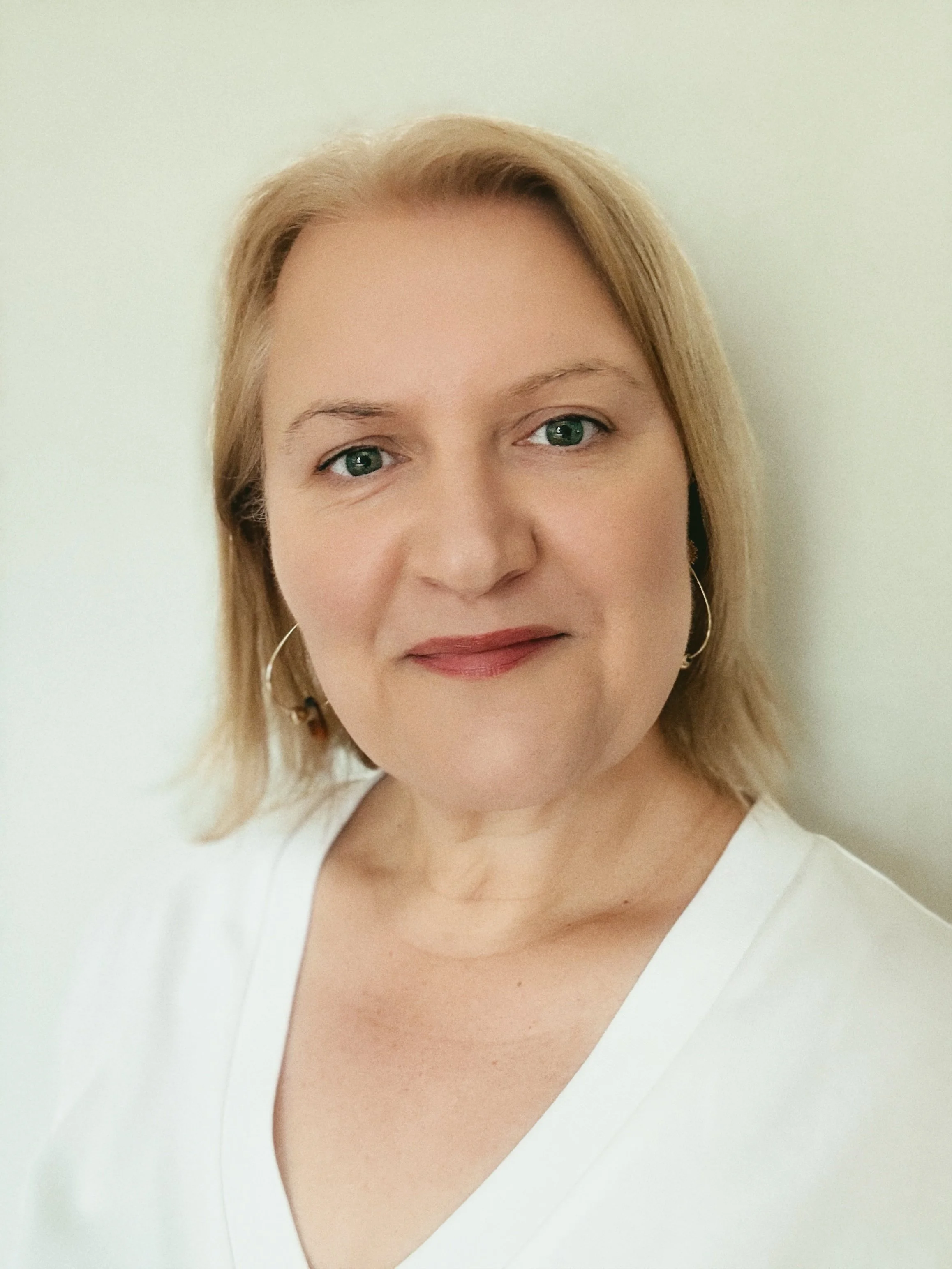 Close-up portrait of a woman with blonde hair, green eyes, wearing a white top and earrings, smiling lightly against a plain background.