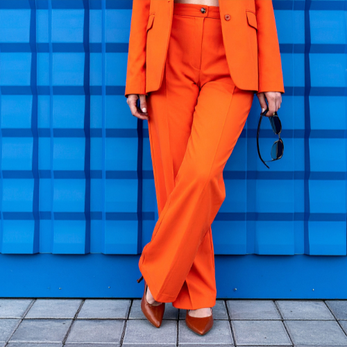 Person wearing an orange pantsuit, holding sunglasses, standing against a blue textured wall.