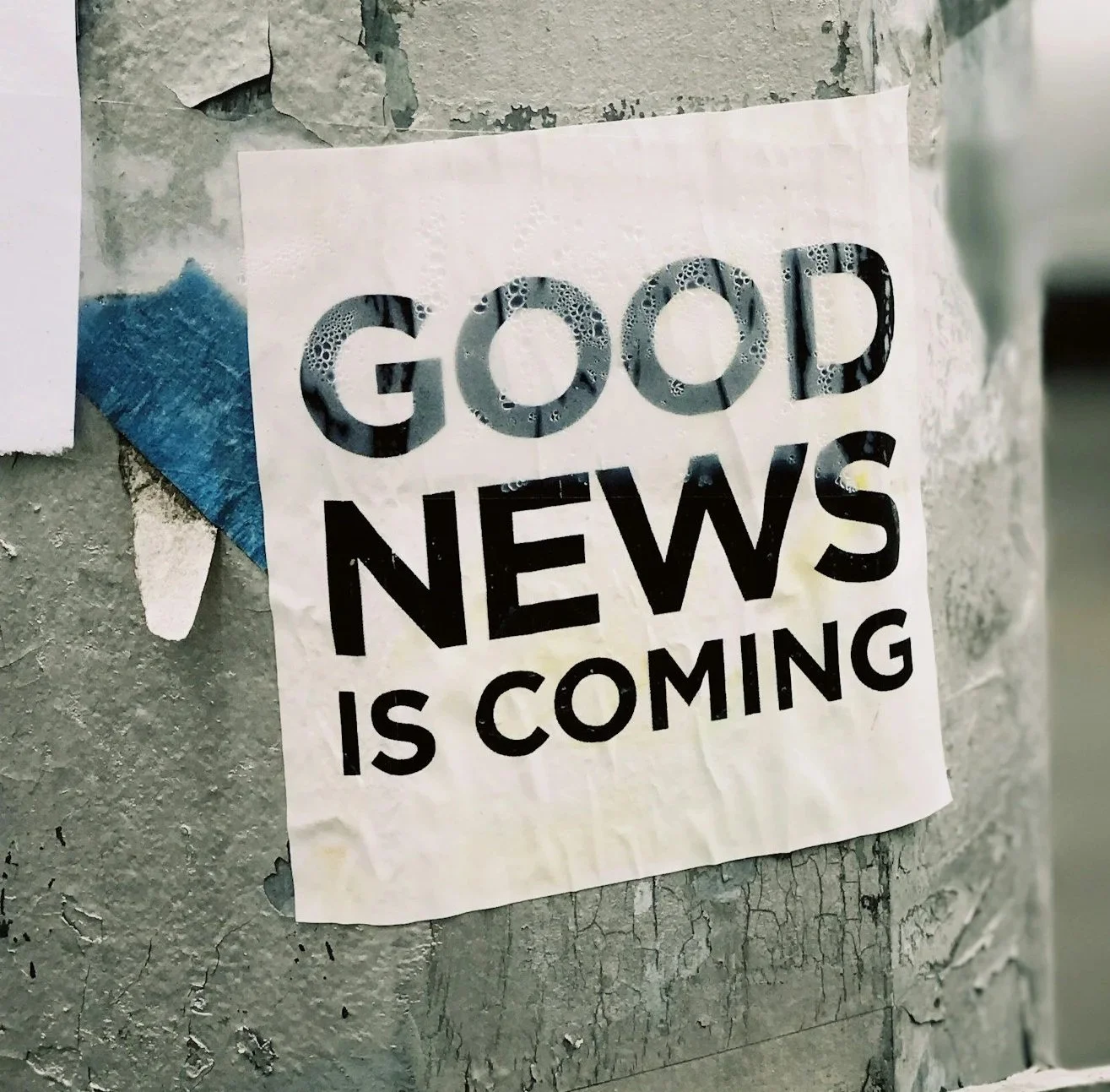 A torn poster on a textured, worn wall that reads, 'GOOD NEWS IS COMING' in large, bold letters, with some soap bubbles covering part of the word 'GOOD.'