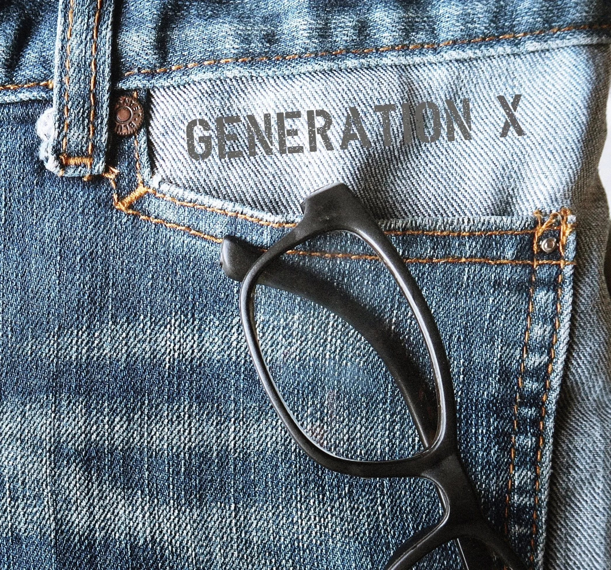 Close-up of a pair of blue denim jeans with a black pair of sunglasses hanging from the belt loop. The jeans have orange stitching and a label that reads 'Generation X'.