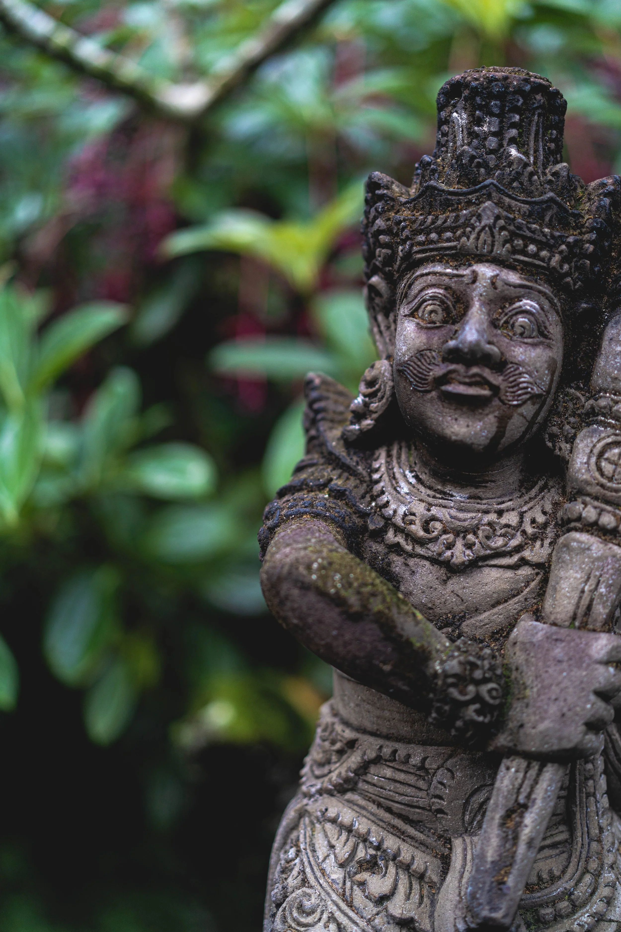Close-up of a stone statue of a warrior or deity with ornate jewelry and a crown, surrounded by lush greenery.