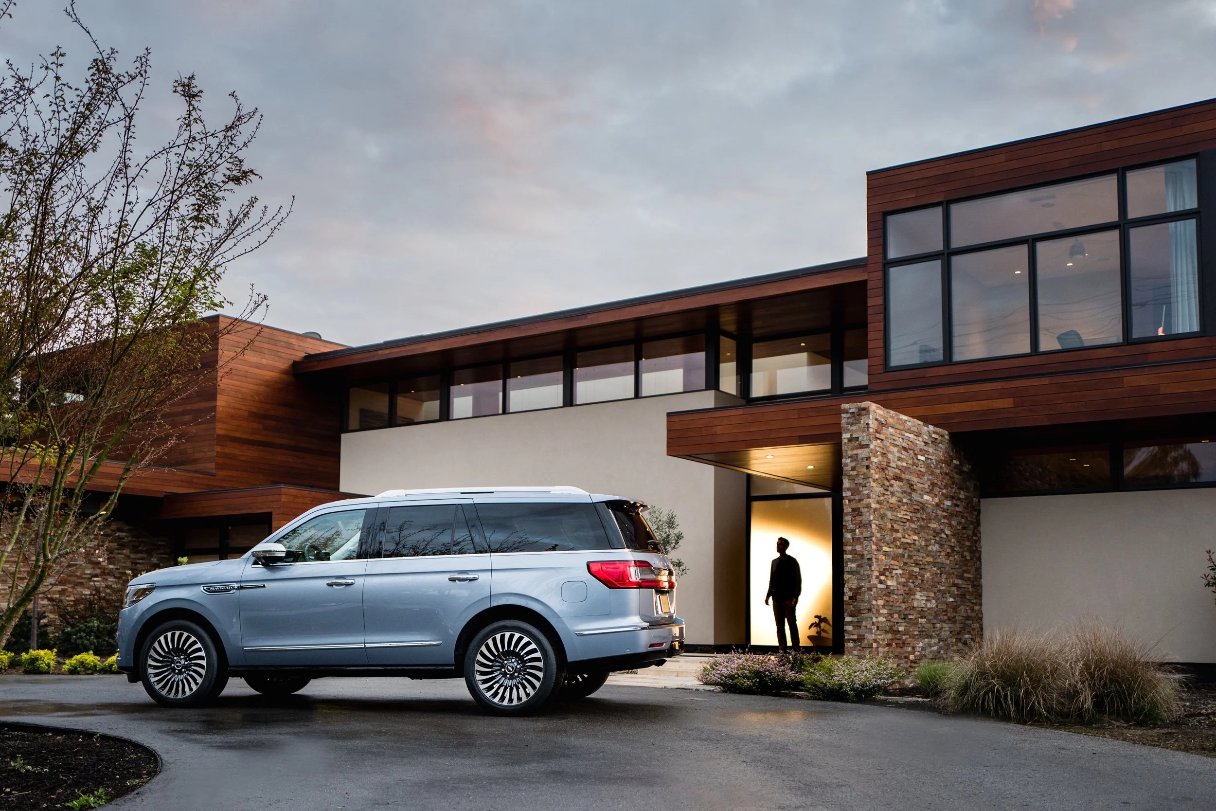 Modern house with wooden and stone exterior, driveway, and a blue SUV parked in front, at dusk. Person standing in the doorway illuminated from inside.