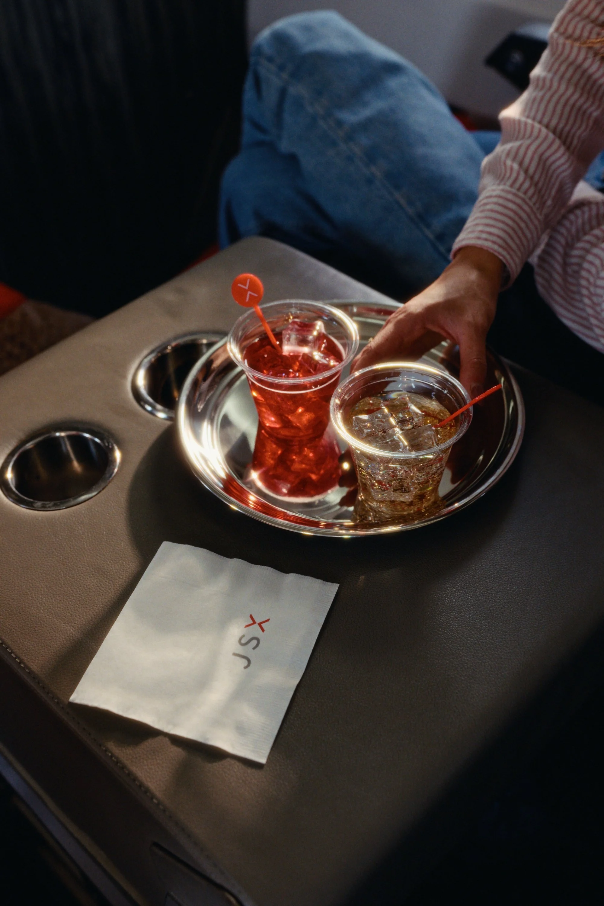 Two drinks on a metal tray, one red and one clear, with ice, on a passenger seat with a person in a striped shirt reaching for one. There are empty cup holders on a table and a napkin with a red logo.