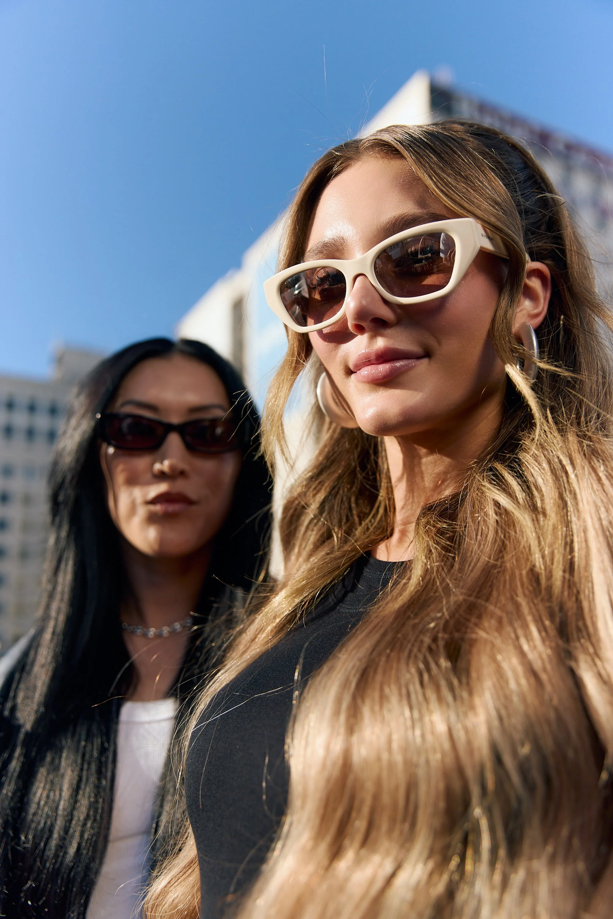 Two women with long hair wearing sunglasses and stylish clothing pose outdoors under a bright blue sky, with city buildings in the background.