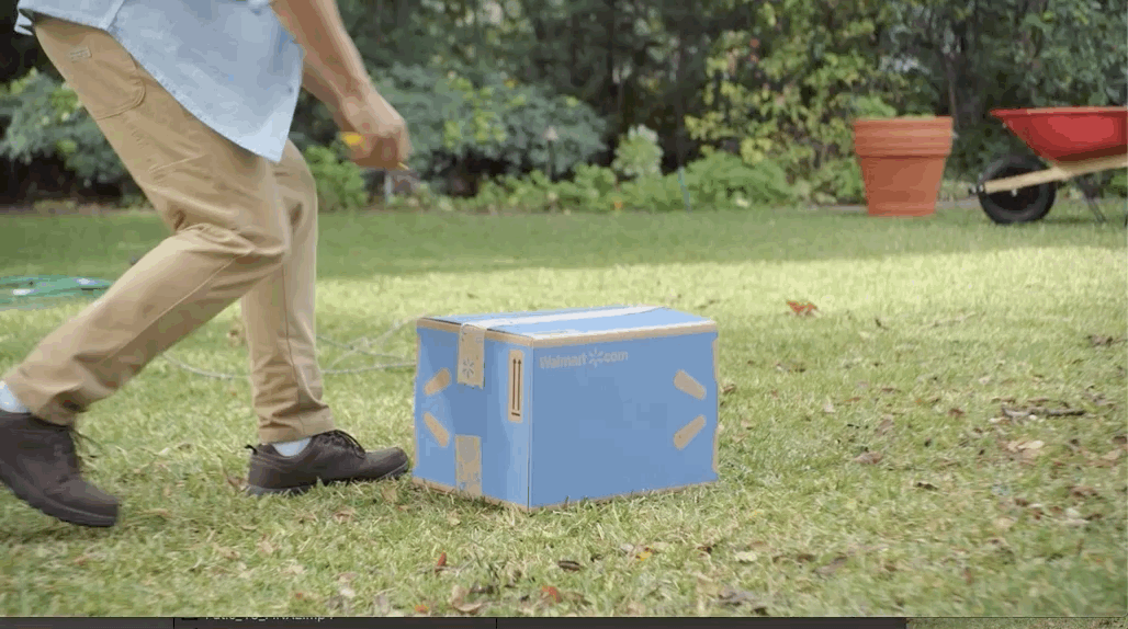 Person wearing khaki pants and black shoes pushing a blue cardboard box on grass in a backyard.