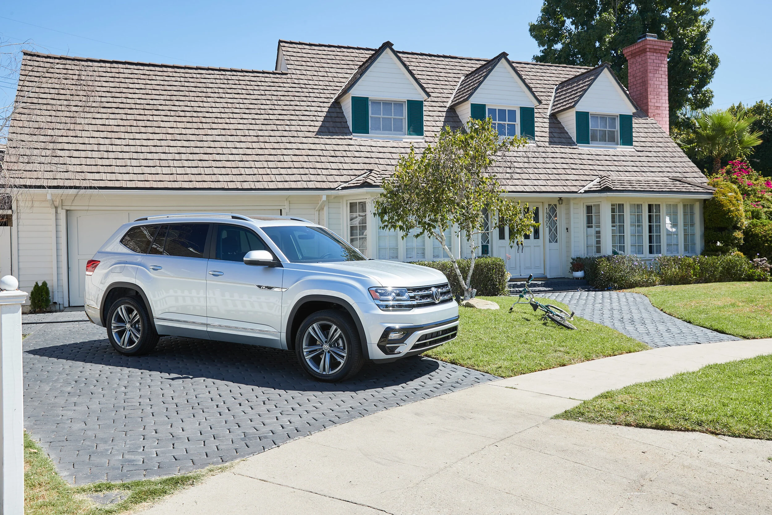 White SUV parked on a stone driveway in front of a white house with green shutters and a bicycle lying on its side on the grass.