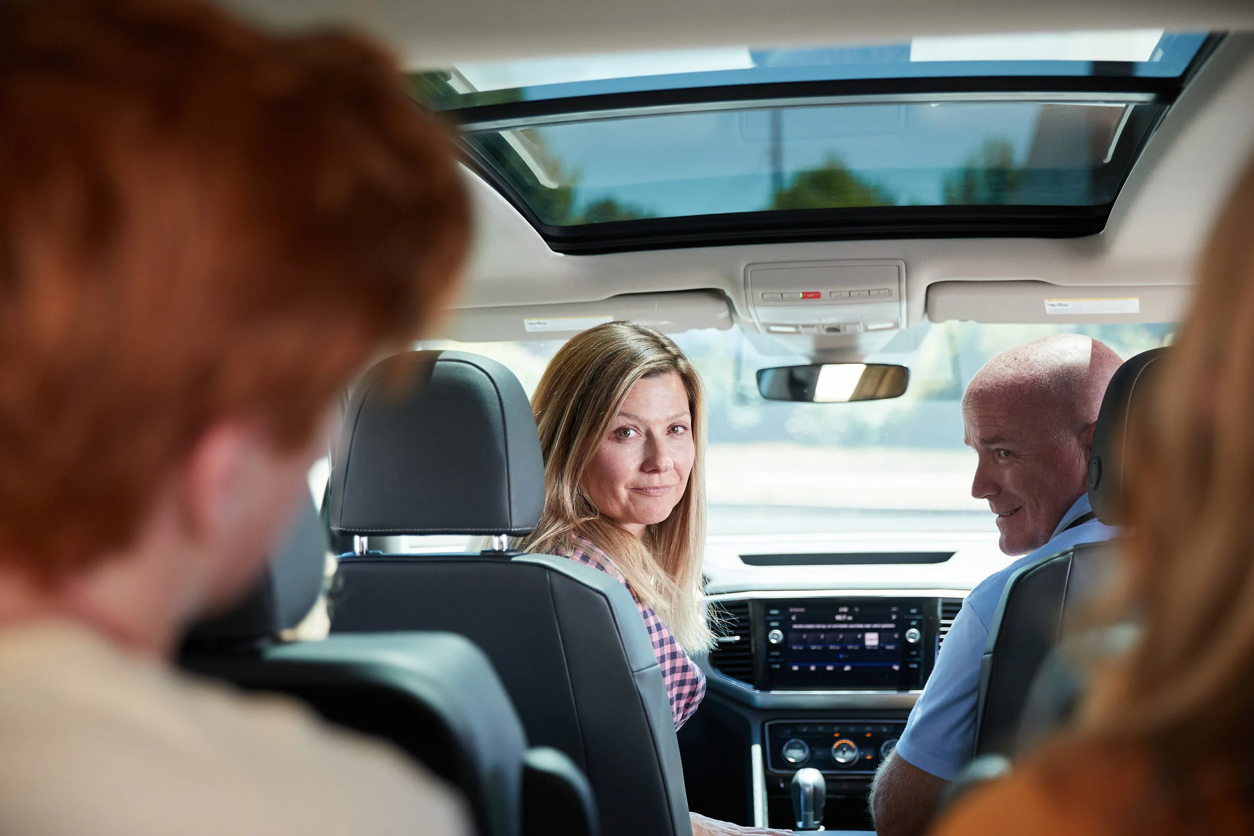 Four people sitting in a car, with one woman and one man in the front seats and two people in the back, the woman is looking towards the camera while the man is looking at her.