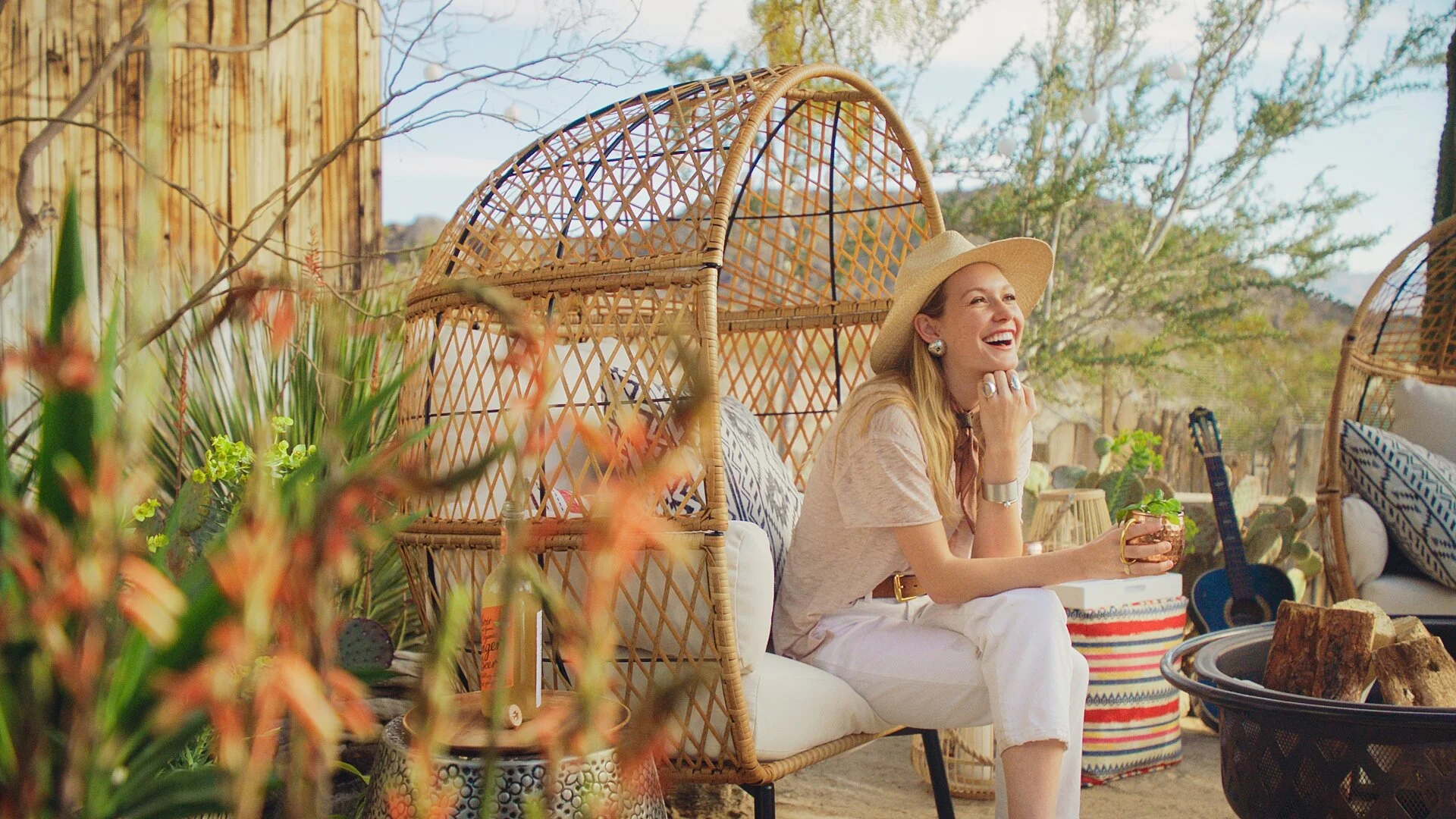 A woman with a wide-brimmed hat sitting outdoors, smiling, with plants, a guitar, and cozy furniture around her.