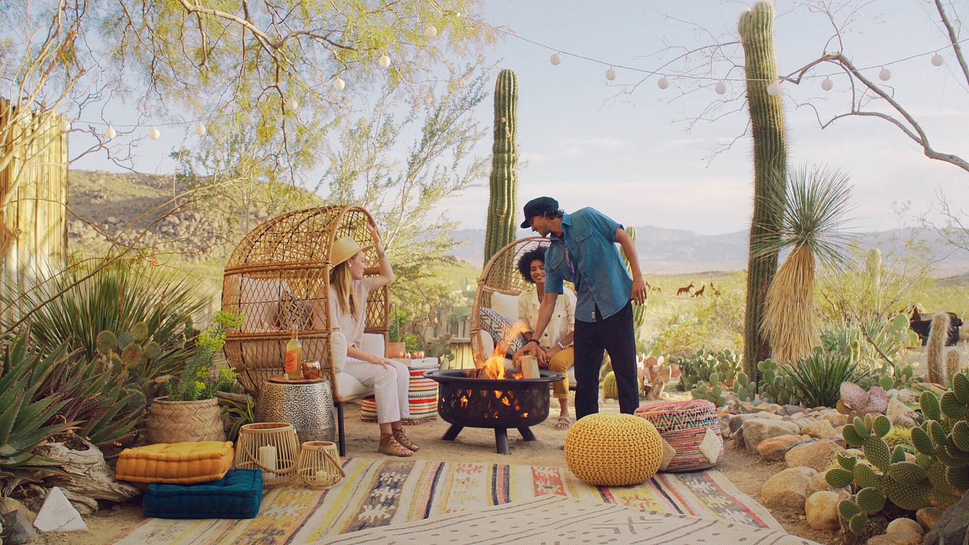 Friends enjoying a barbecue around a fire pit in a desert landscape with cacti and mountains, sitting on bohemian furniture and rugs.