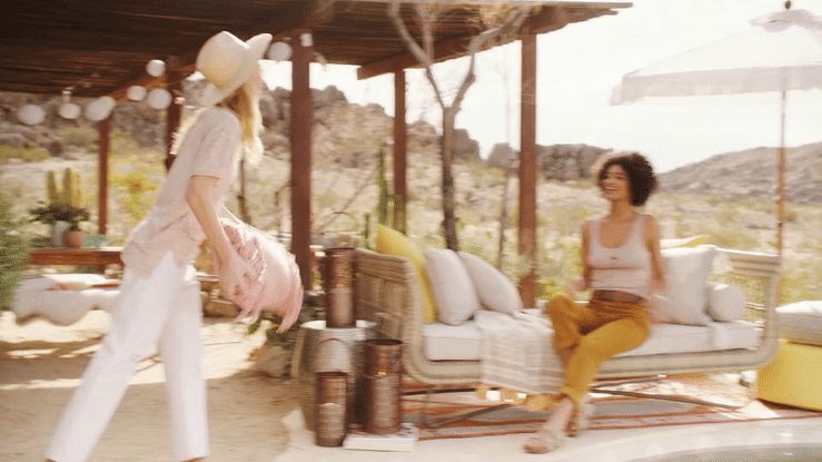 Two women enjoying conversation outdoors under a covered patio, with one sitting on a sofa and the other approaching, in a sunny, desert-like setting.