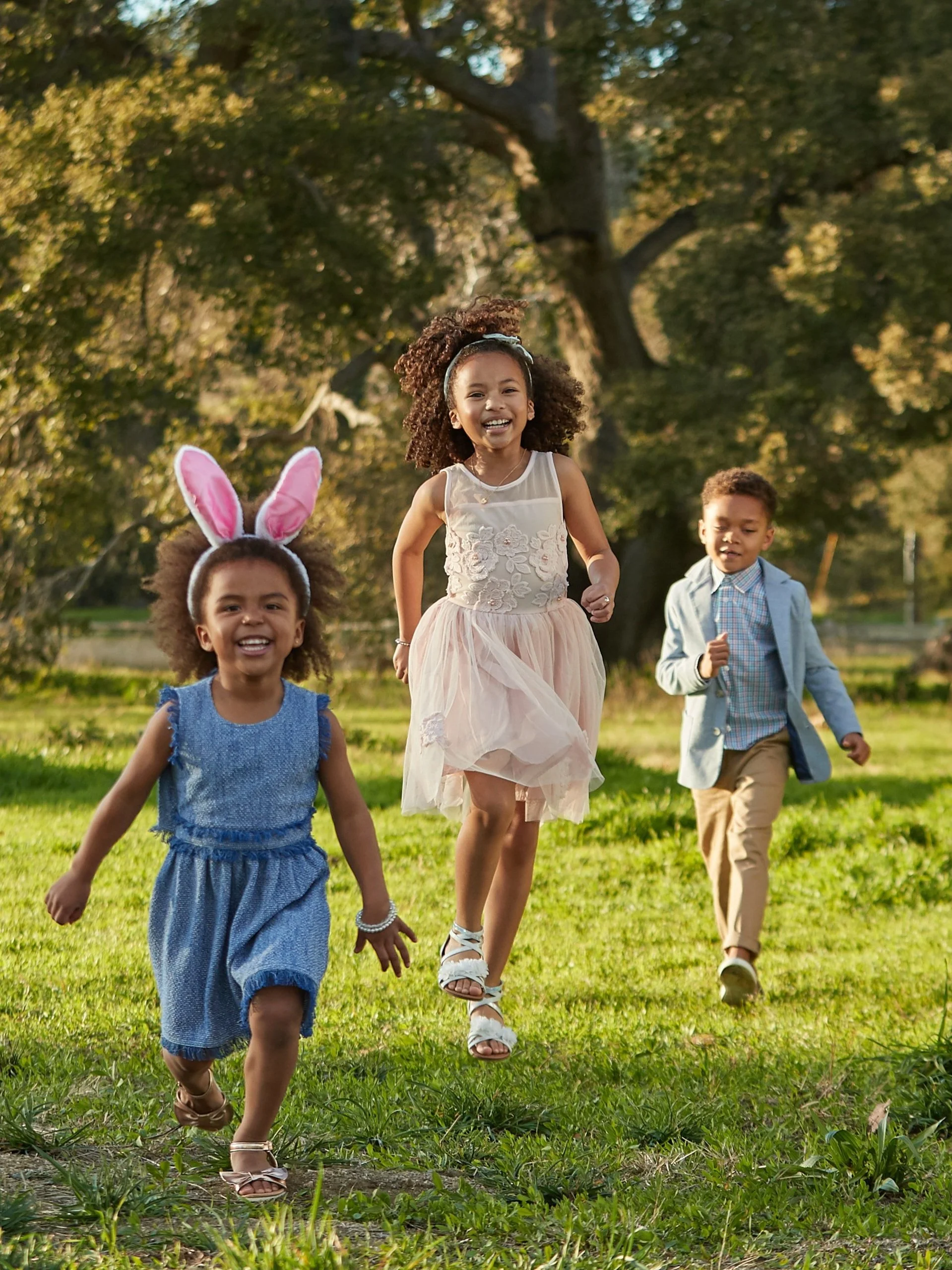 Three children running and playing outdoors on a sunny day, dressed in festive clothes, with one girl wearing bunny ears.