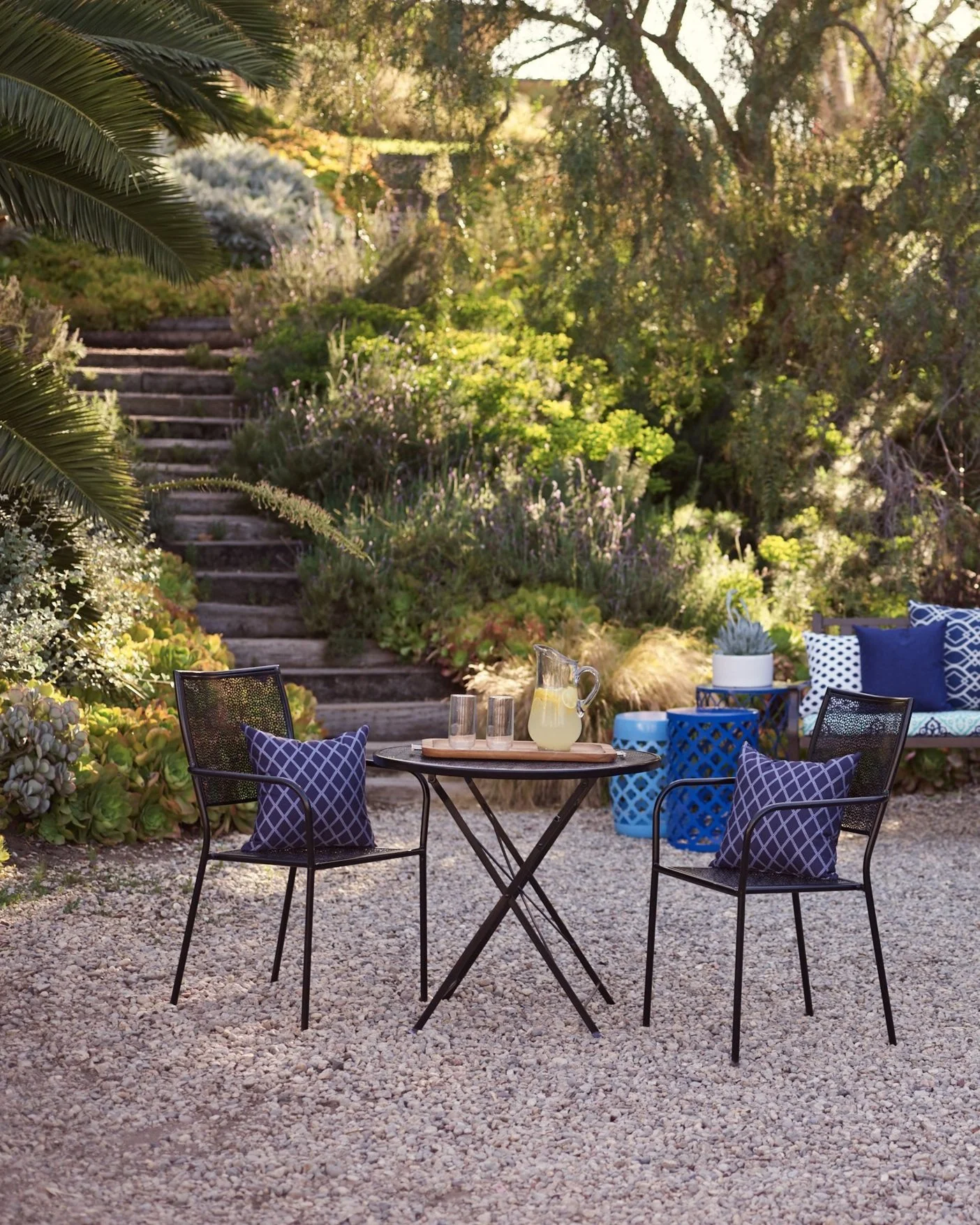 A cozy outdoor patio scene with a small round table, two metal chairs with blue pillows, and a wooden bench with blue cushions and patterned pillows, surrounded by greenery and a gravel ground.