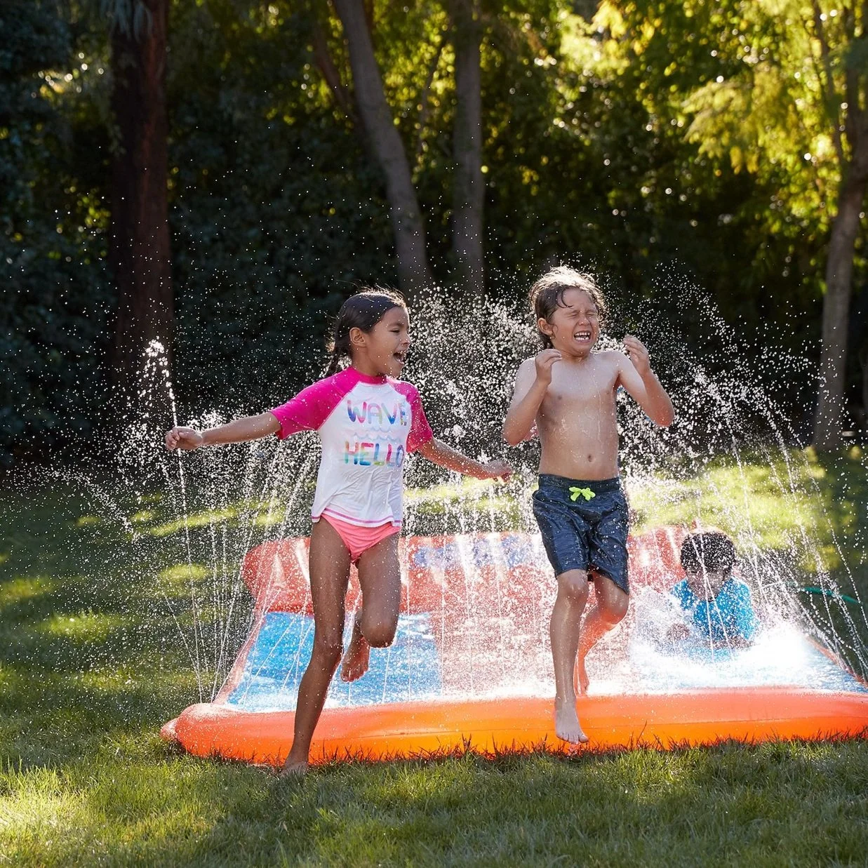Two children playing and running in an inflatable water pool in a backyard on a sunny day, with a third child in the background.