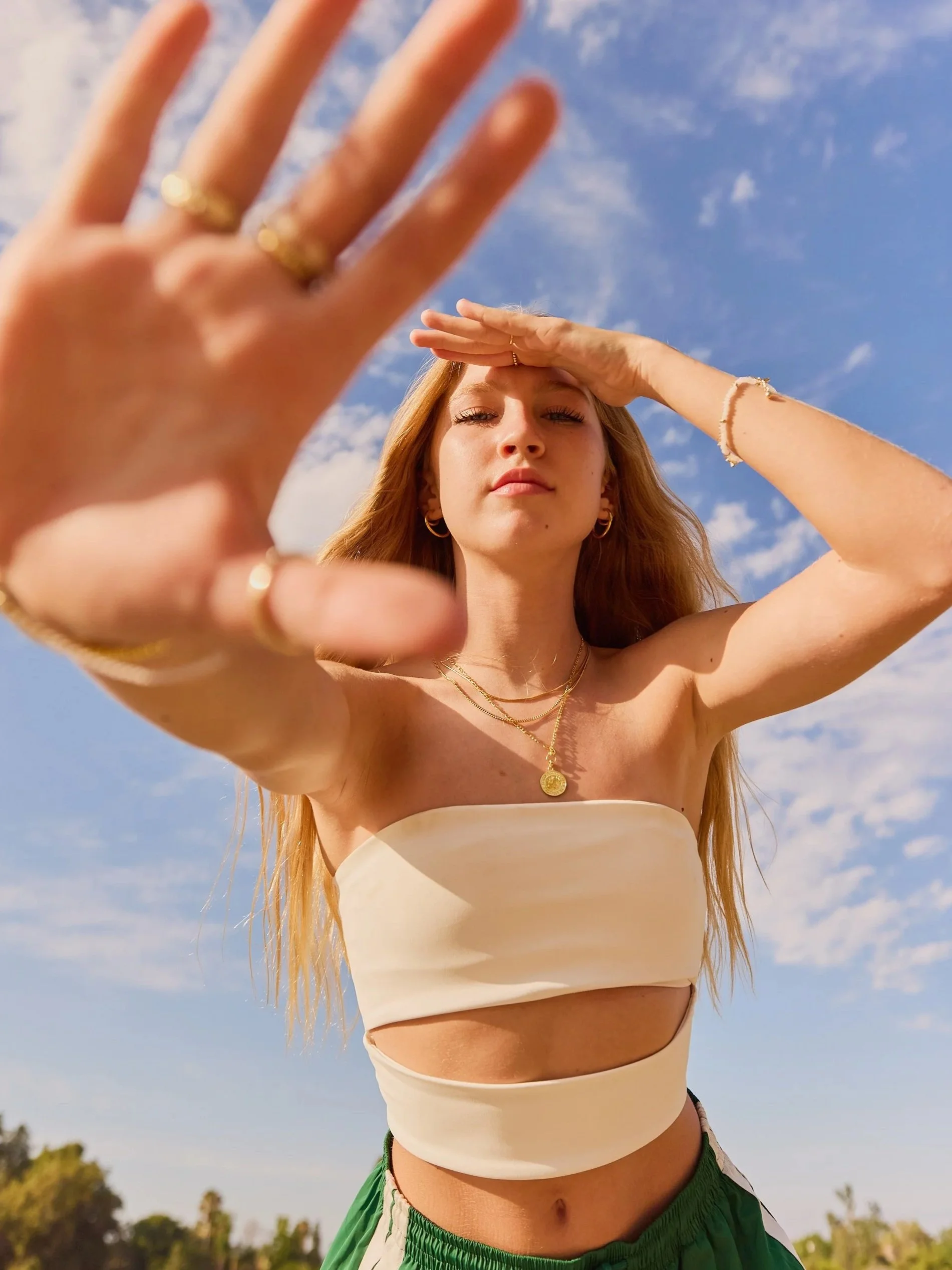 Young woman with long red hair, wearing jewelry and a cream-colored strapless top, shielding her eyes from the sun with her hand, outdoors during daytime with a blue sky and clouds.
