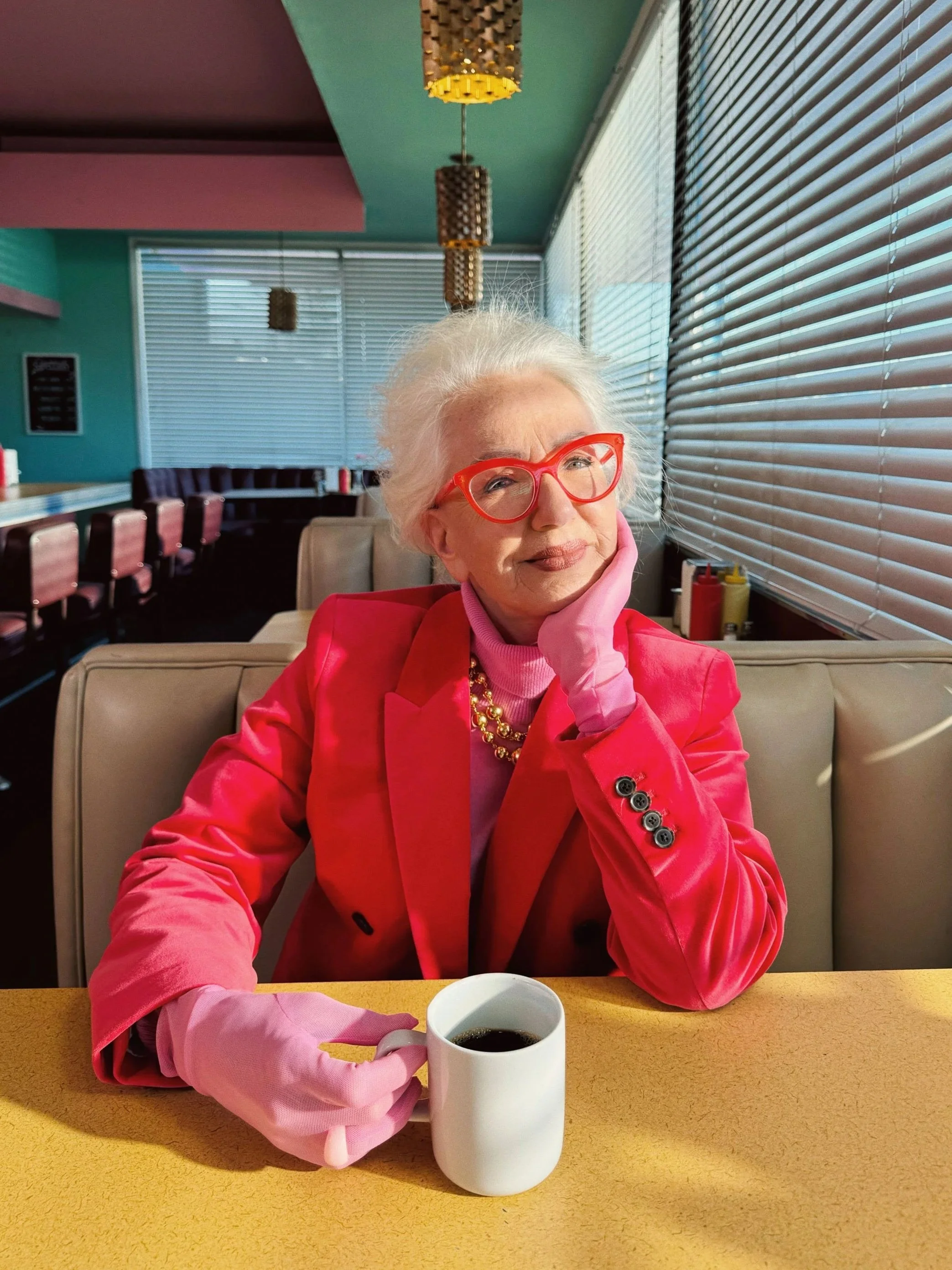 An elderly woman with white hair, red glasses, and a bright pink blazer sitting in a retro diner with a yellow table, holding a white coffee mug. She is wearing pink gloves, a pink turtleneck, and a gold necklace, resting her face on her hand and loo