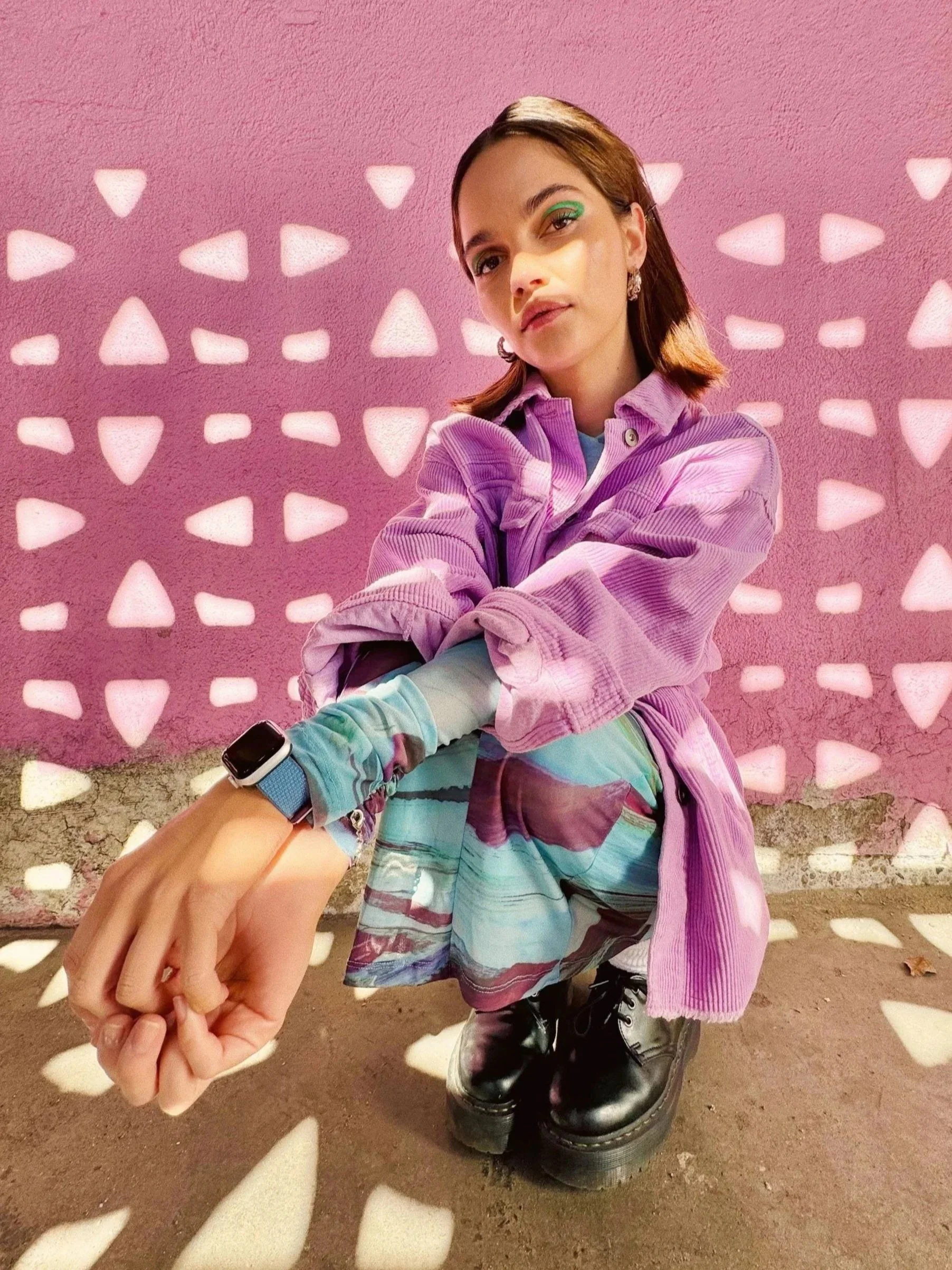 A young woman with light skin tone and shoulder-length brown hair wearing a purple jacket, a colorful skirt, and black platform boots, squatting in front of a pink wall with white triangular cutouts, looking at the camera.