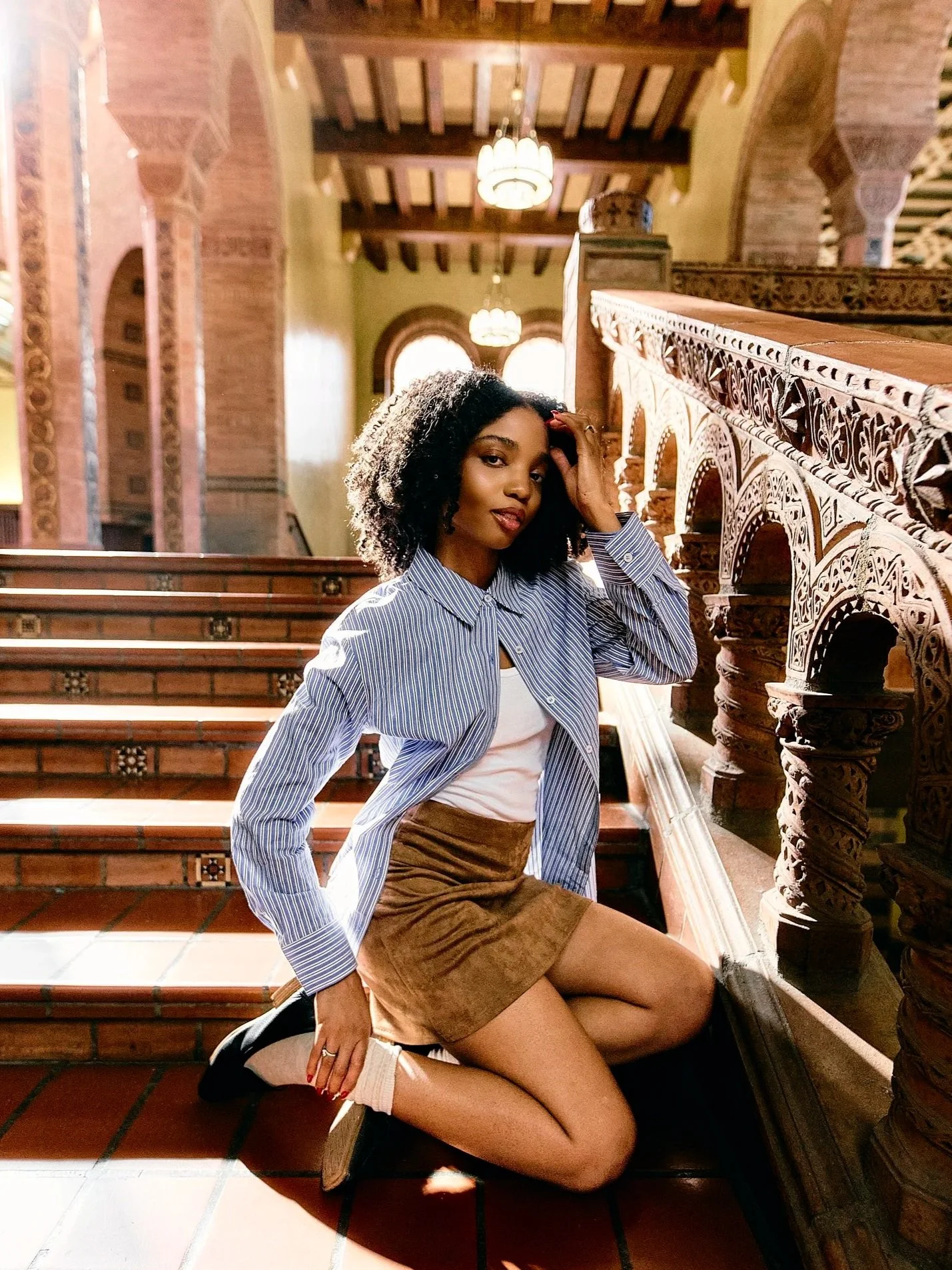 A young woman with curly black hair kneeling on the staircase of a historic building with ornate wooden railings, dressed in a striped blue shirt, white top, and brown skirt, with sunlight streaming through arched windows in the background.