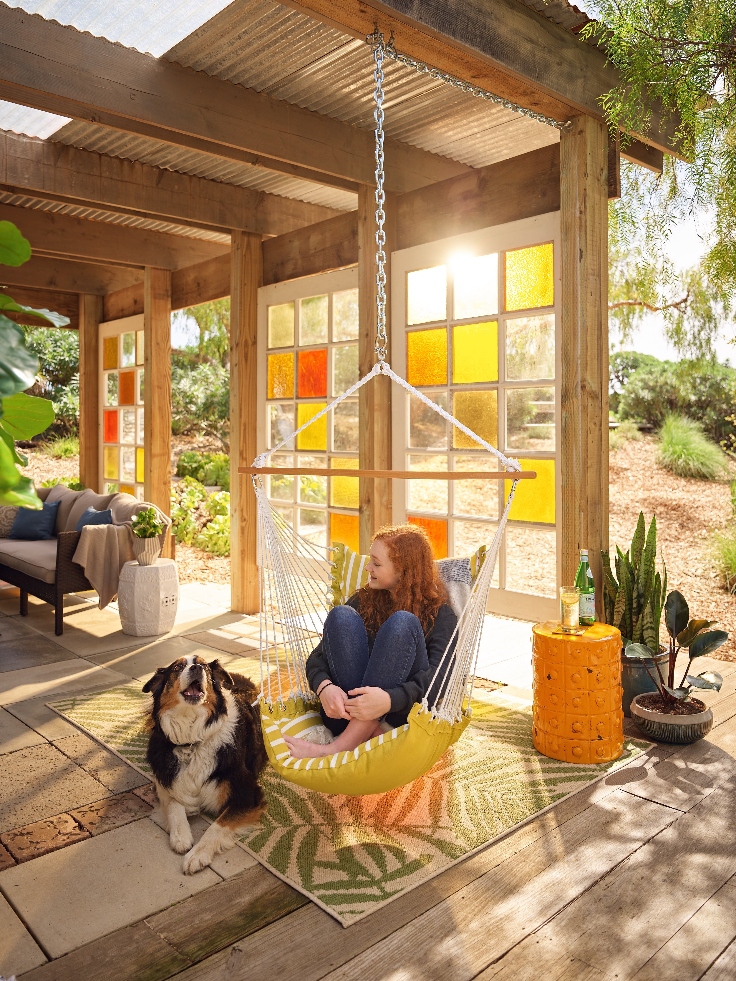 A woman with red hair sitting on a yellow hammock chair with a black and white dog beside her inside a sunlit outdoor patio with colorful stained glass windows, potted plants, and outdoor furniture.
