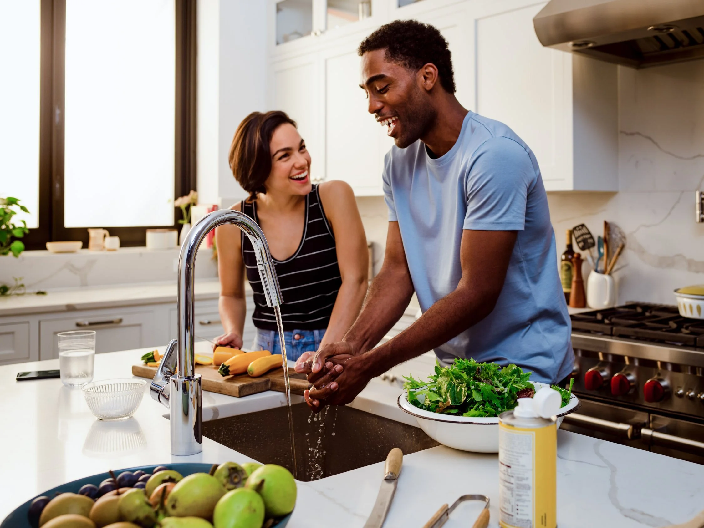 A man is washing vegetables at a kitchen sink, while a woman watches and laughs, in a bright modern kitchen.