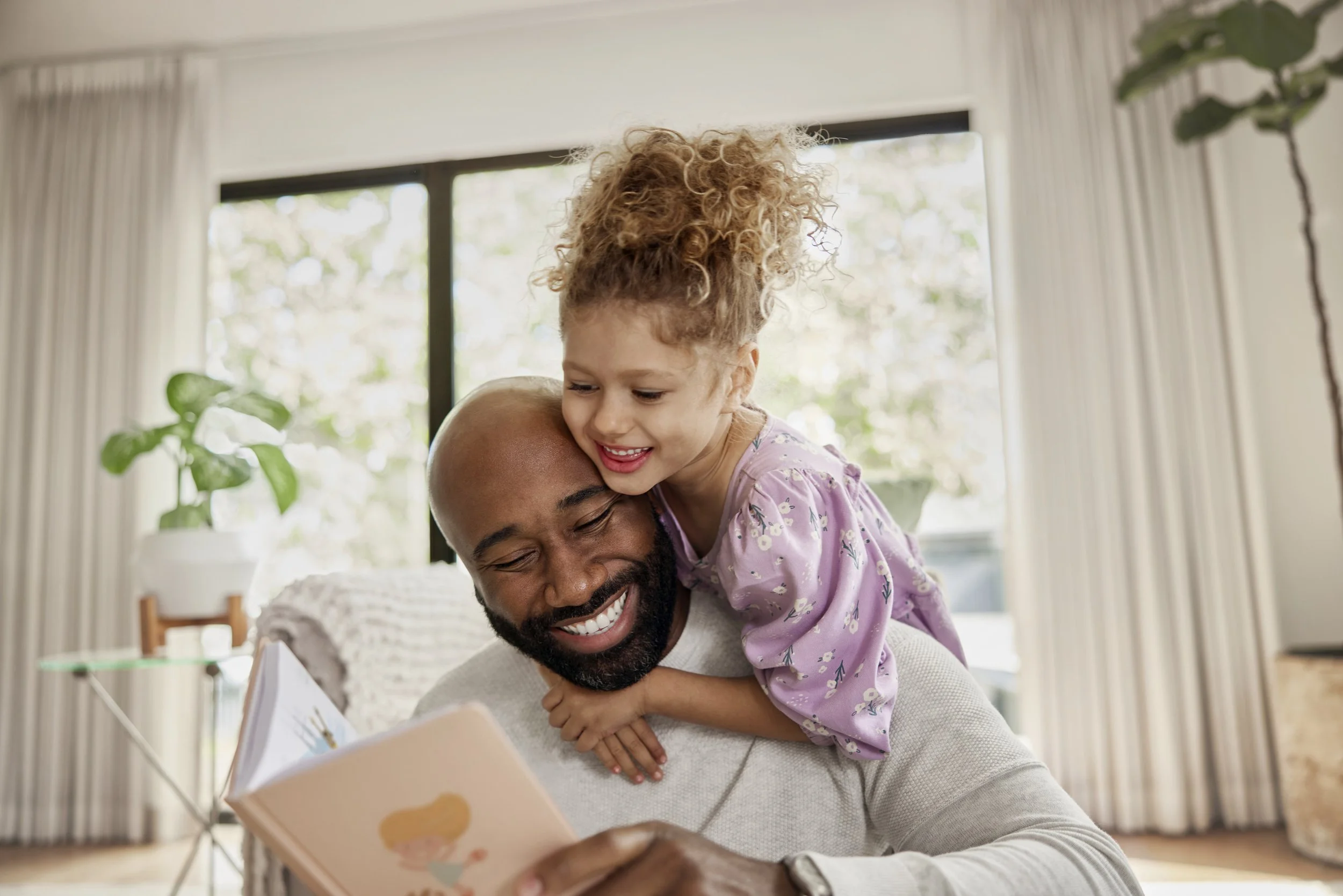 A man and young girl sharing a joyful moment while looking at a book together in a bright living room.