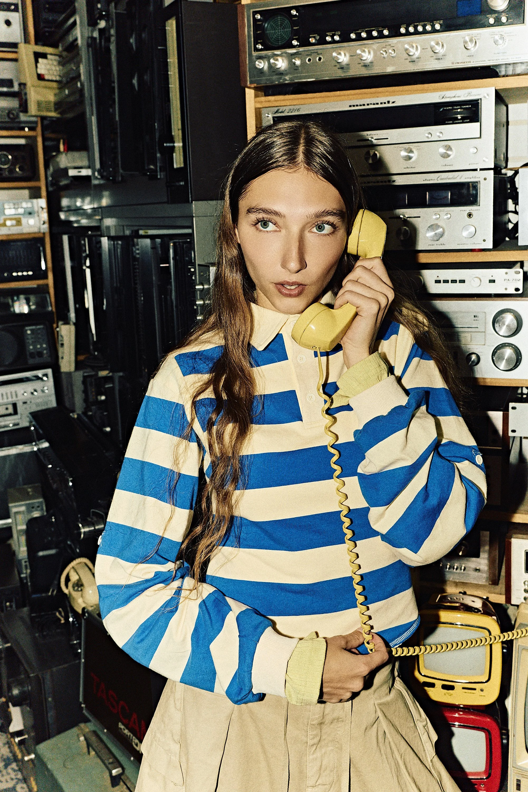 A young woman with long wavy hair, wearing a blue and cream striped shirt and beige skirt, is holding a yellow telephone receiver to her ear. She is standing in a room filled with vintage audio equipment and electronics.