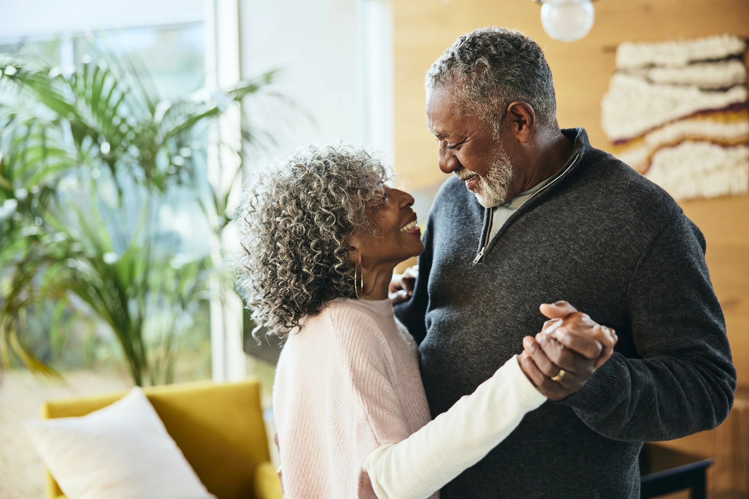 An older couple dancing together in a cozy, well-lit living room with a large window, a yellow armchair, and green plants in the background. They are smiling and looking at each other lovingly.