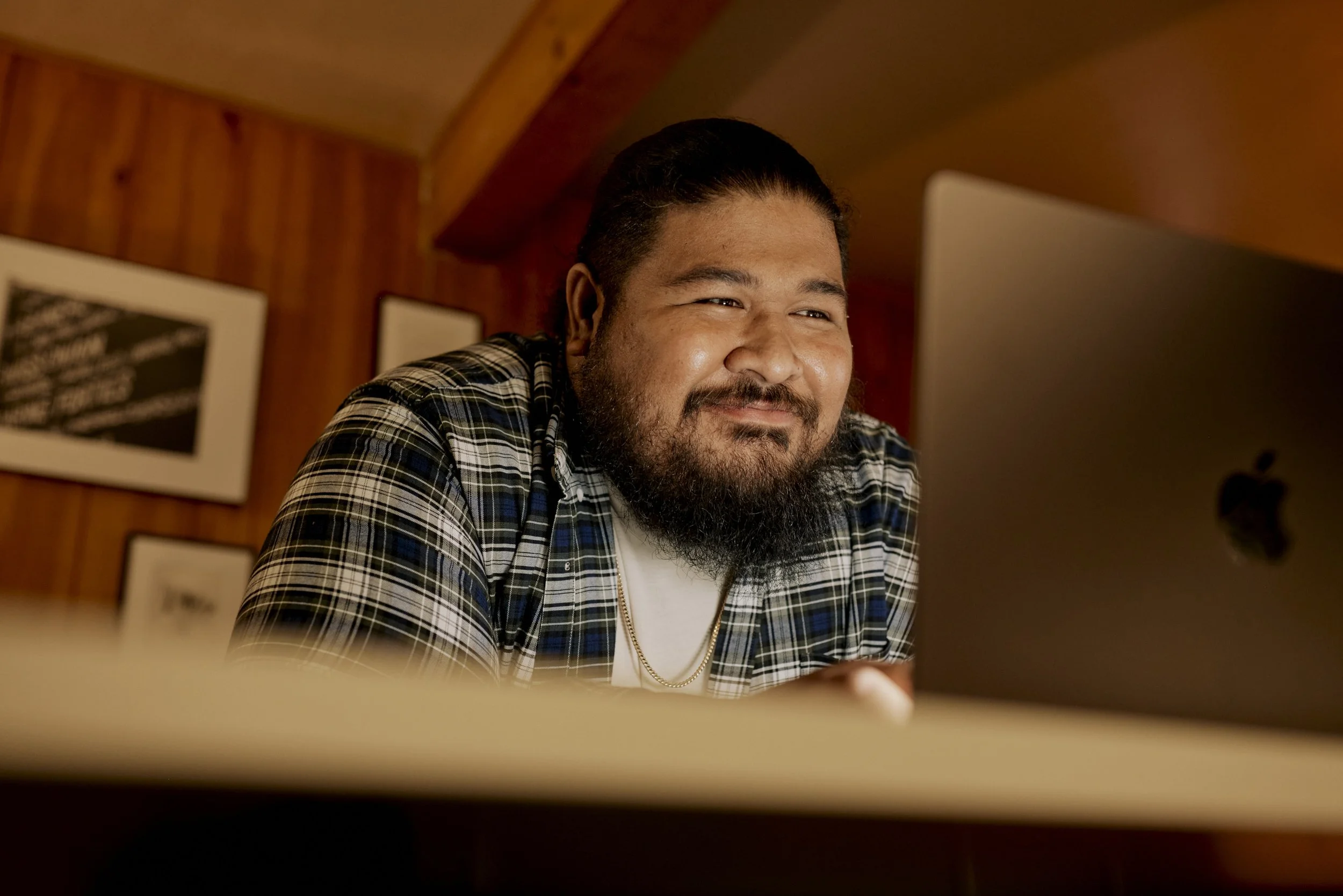 A man with a beard and long hair tied back, wearing a checkered shirt and a gold chain, smiling while looking at a computer screen in a wooden-paneled room.