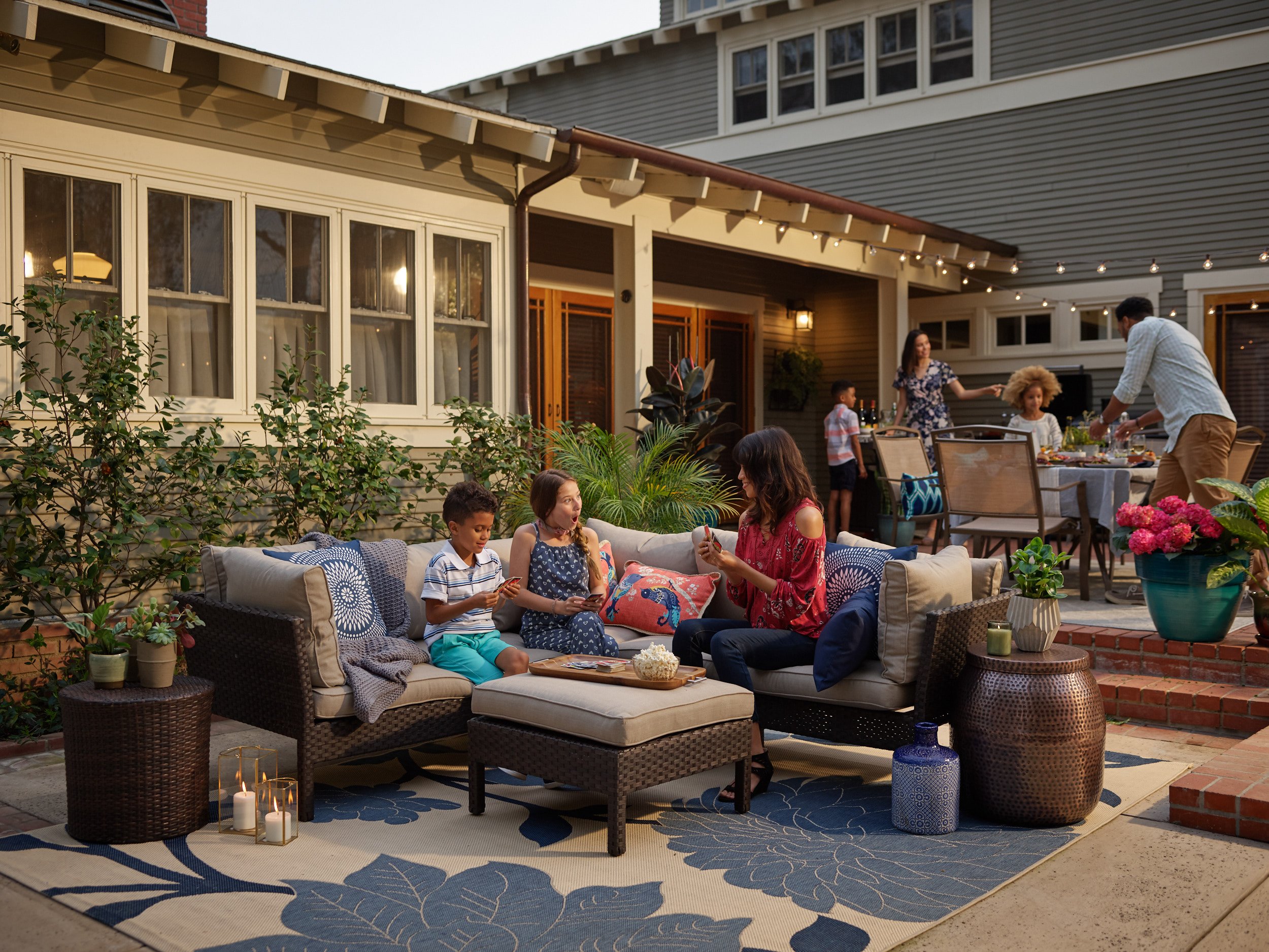 Family gathering on a backyard patio during the evening, with children and adults enjoying food and conversations. Cushioned seating area with decorative pillows, a table with candles, plants, and a dining table in the background with food and drinks