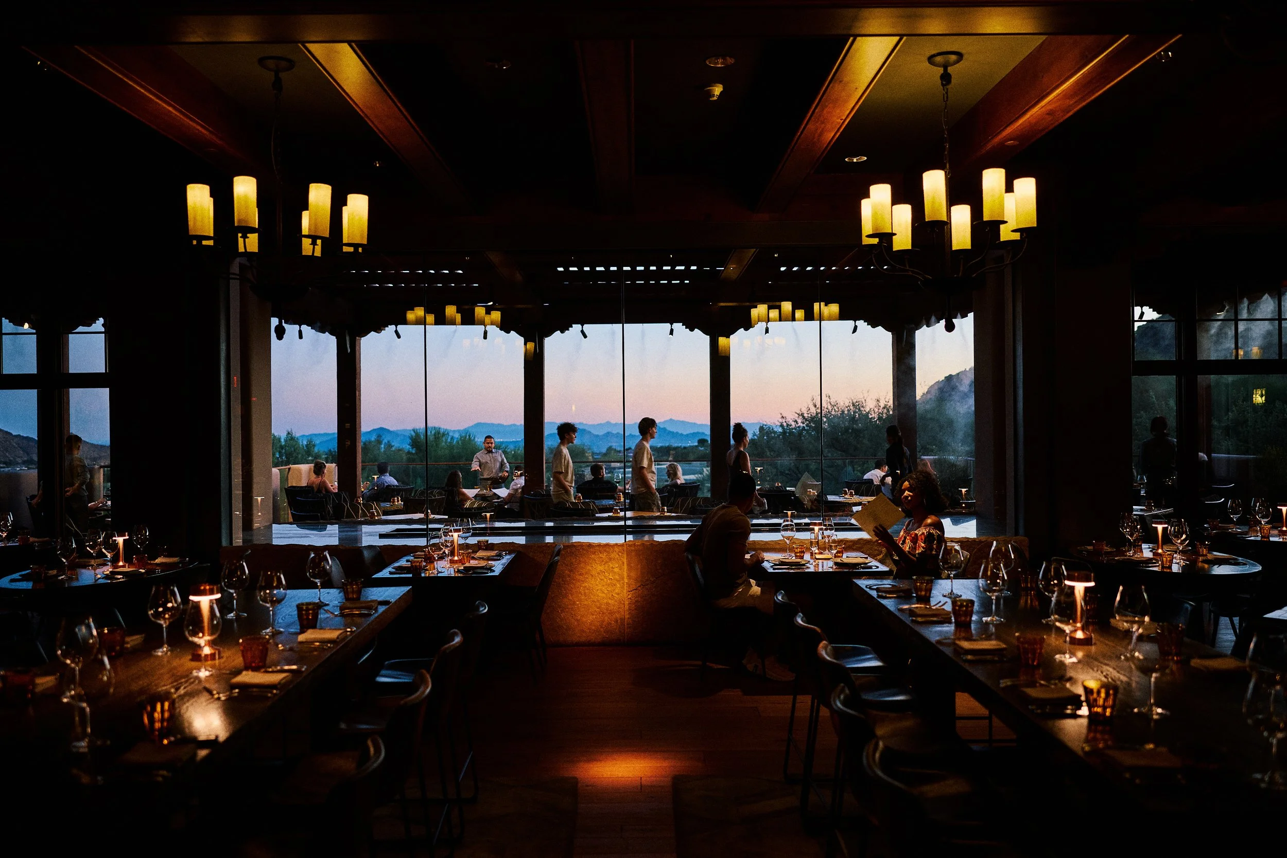 Elegantly set restaurant with dim lighting, large windows showing a scenic mountain view at sunset, and several people dining and servers attending to guests inside.