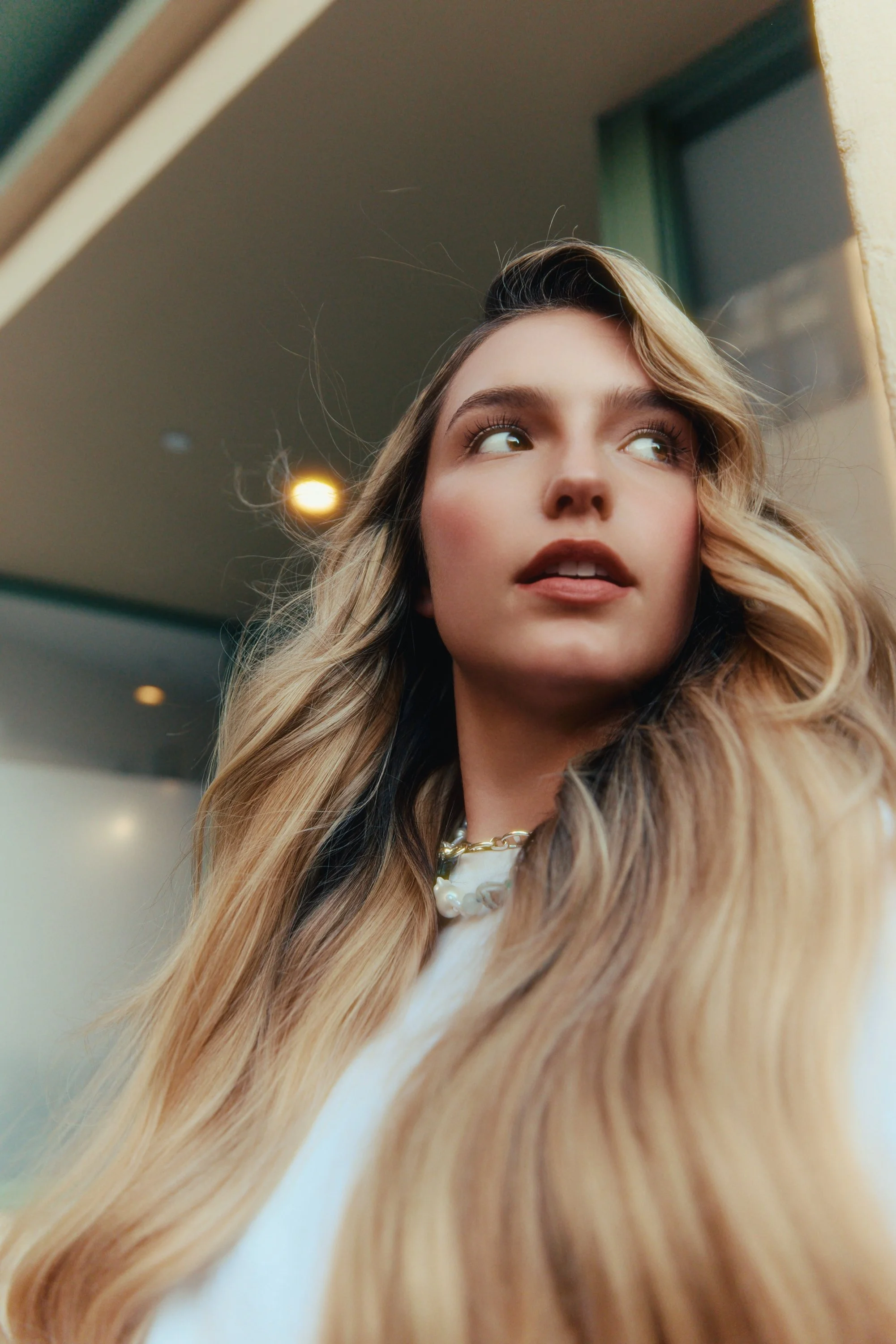 Close-up of a young woman with long, wavy blonde hair looking to the side indoors.