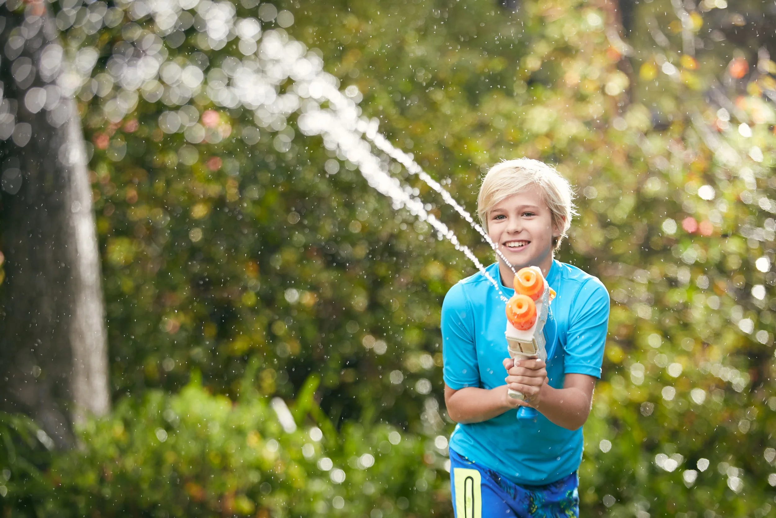 A young boy with blond hair wearing a blue shirt and blue shorts, holding a water gun, smiling while playing outdoors in a sunny, lush green backyard.